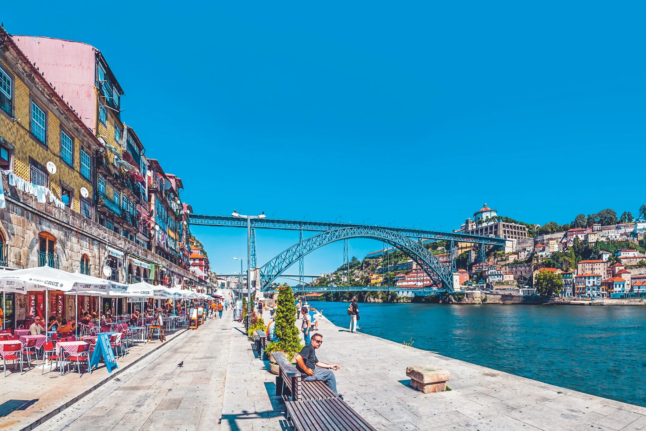 A film and photo location in Portugal from walnut films, People sitting and walking along a riverside promenade with cafe tables and umbrellas, colorful buildings on a hill, a large suspension bridge, and a clear blue sky in Porto, Portugal.