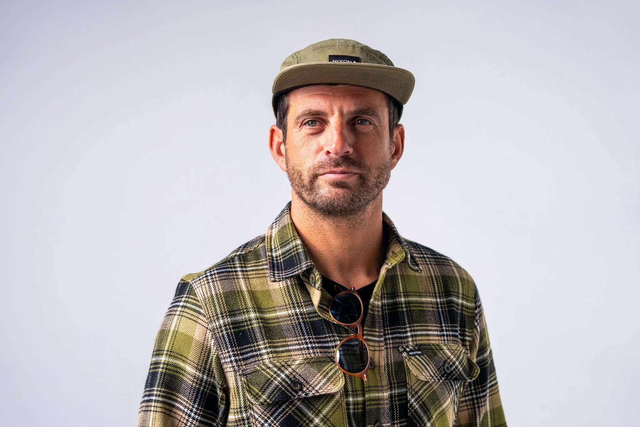A producer from walnut films, wearing a green and yellow plaid shirt, a tan cap, and sunglasses hanging from his shirt, standing against a plain light background.