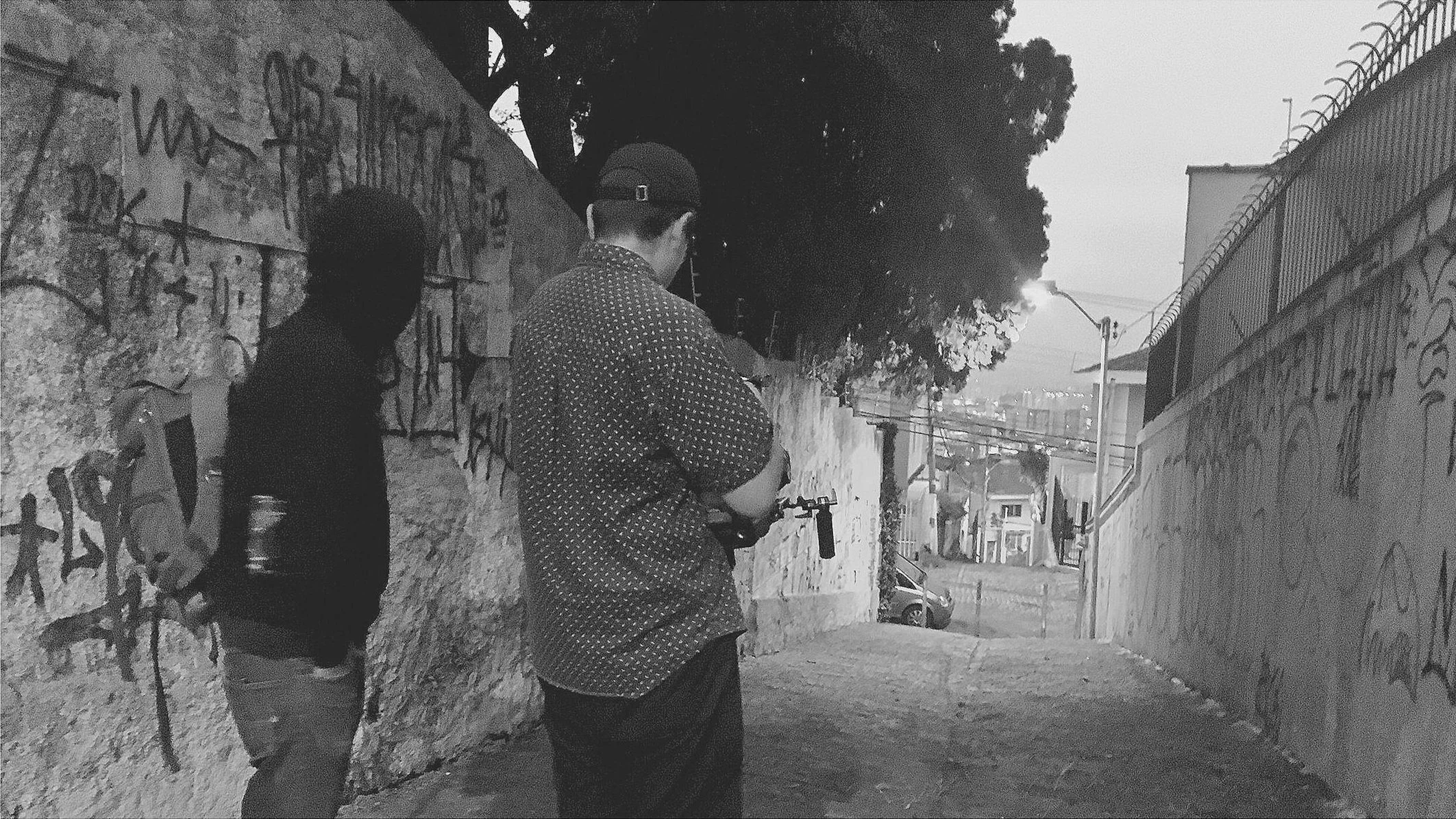 Two individuals standing on an urban sidewalk near a graffiti-covered concrete wall, with trees and buildings in the background, during dusk.