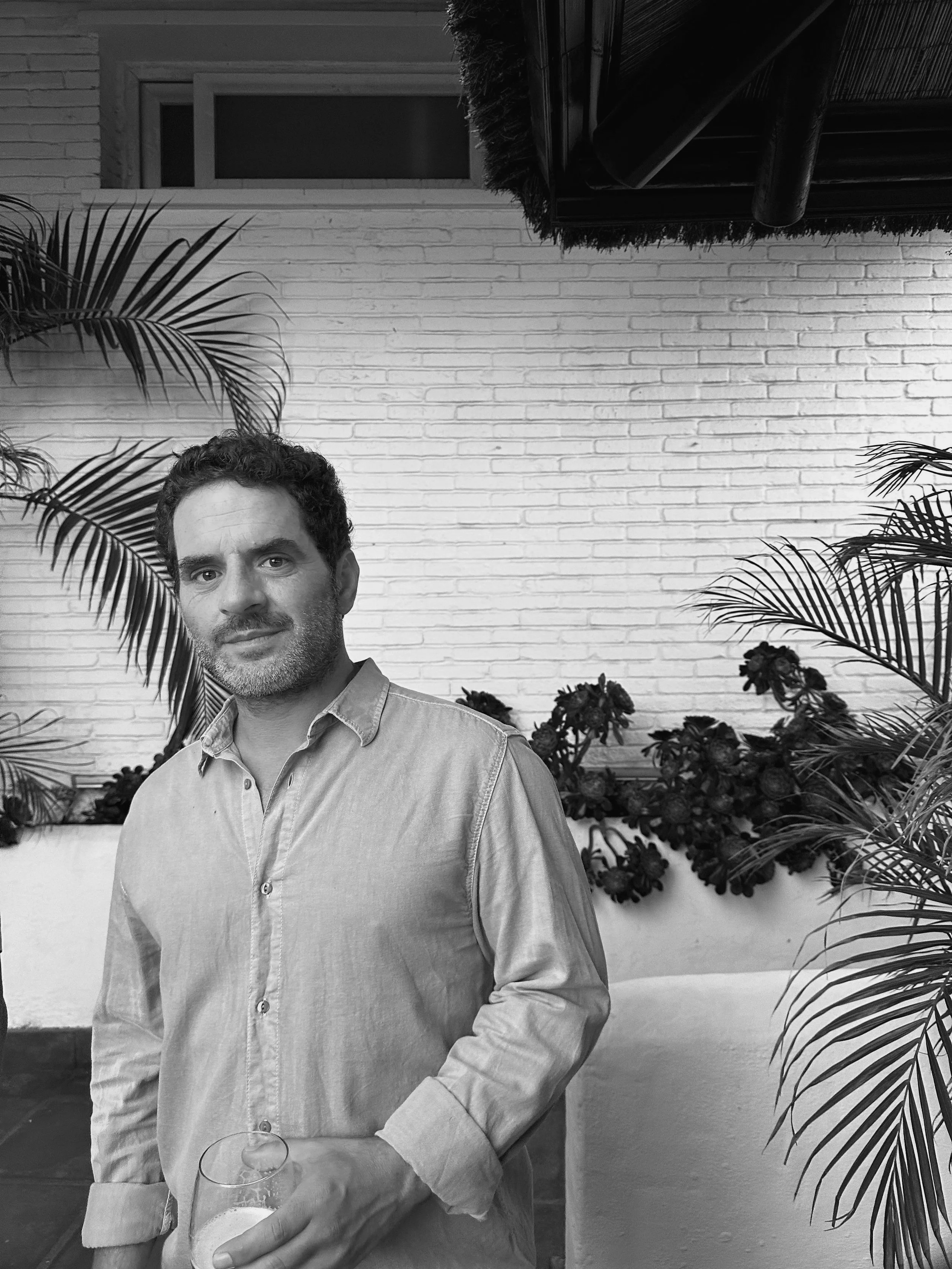 A man with dark, curly hair and a beard, wearing a light-colored button-up shirt, holding a glass of wine in his right hand, standing in front of a bathroom or living room with a white brick wall, a window, and tropical plants in the background.
