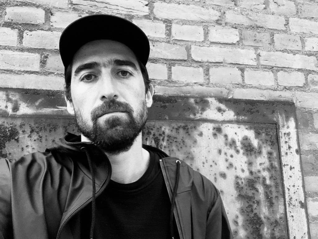 A young man with a beard and mustache wearing a black cap and jacket, taking a selfie in front of a brick wall and a rusty metal panel.