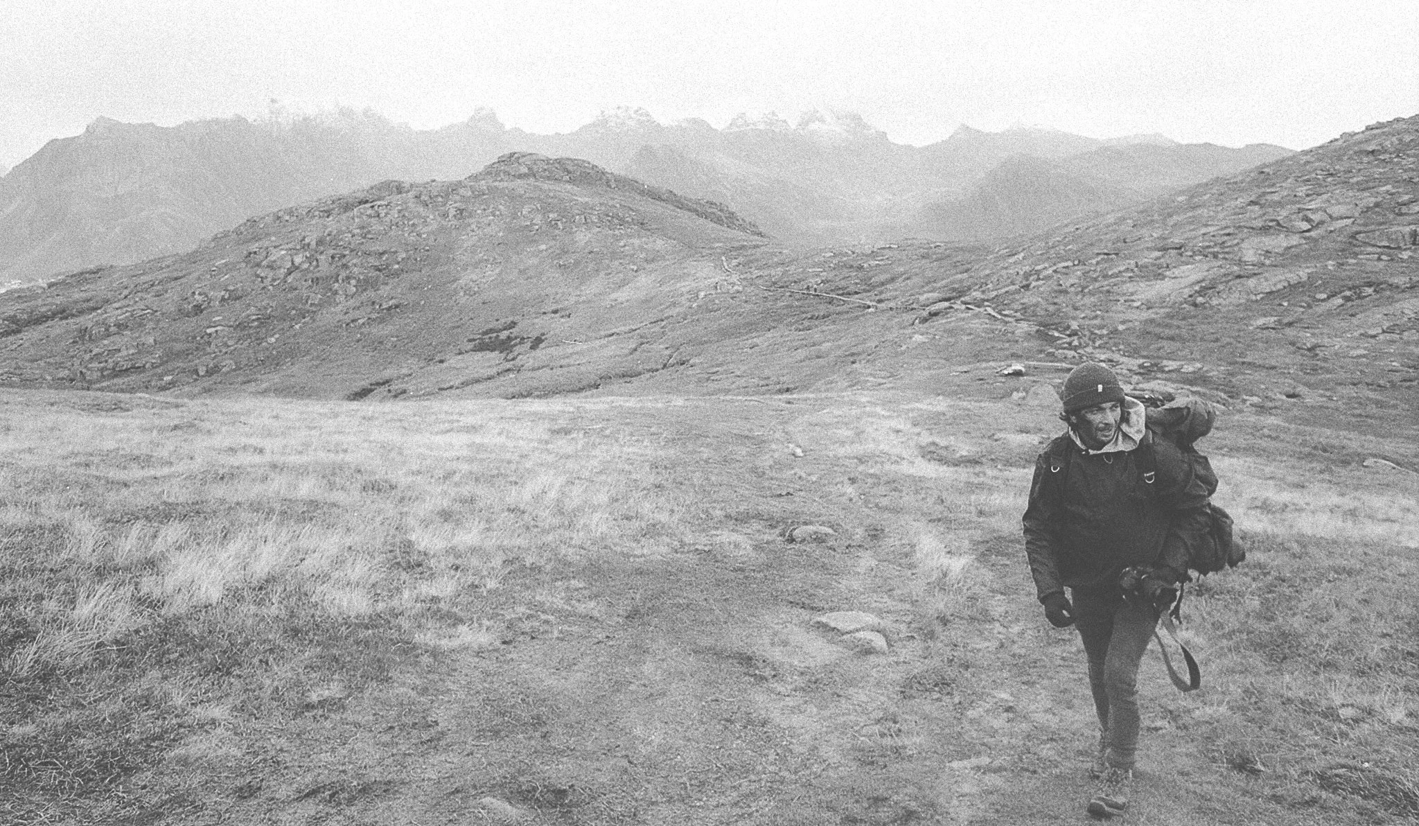 A person in outdoor clothing and a helmet is hiking in a mountainous landscape with grassy terrain, rocky hills, and distant peaks.