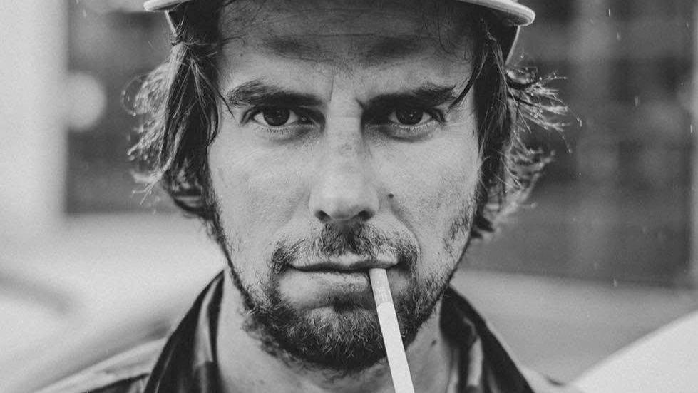 Close-up of a man with unkempt hair and a beard, holding a cigarette in his mouth, wearing a cap.