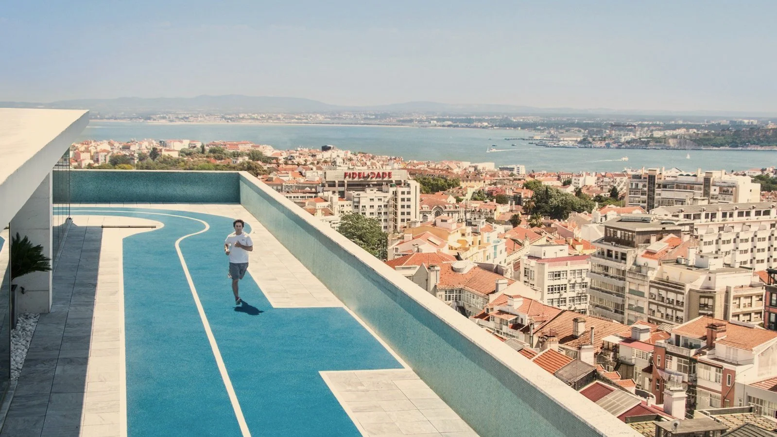 A man jogging on a rooftop running track overlooking a cityscape with red-tiled roofs, buildings, and a river in the background.
