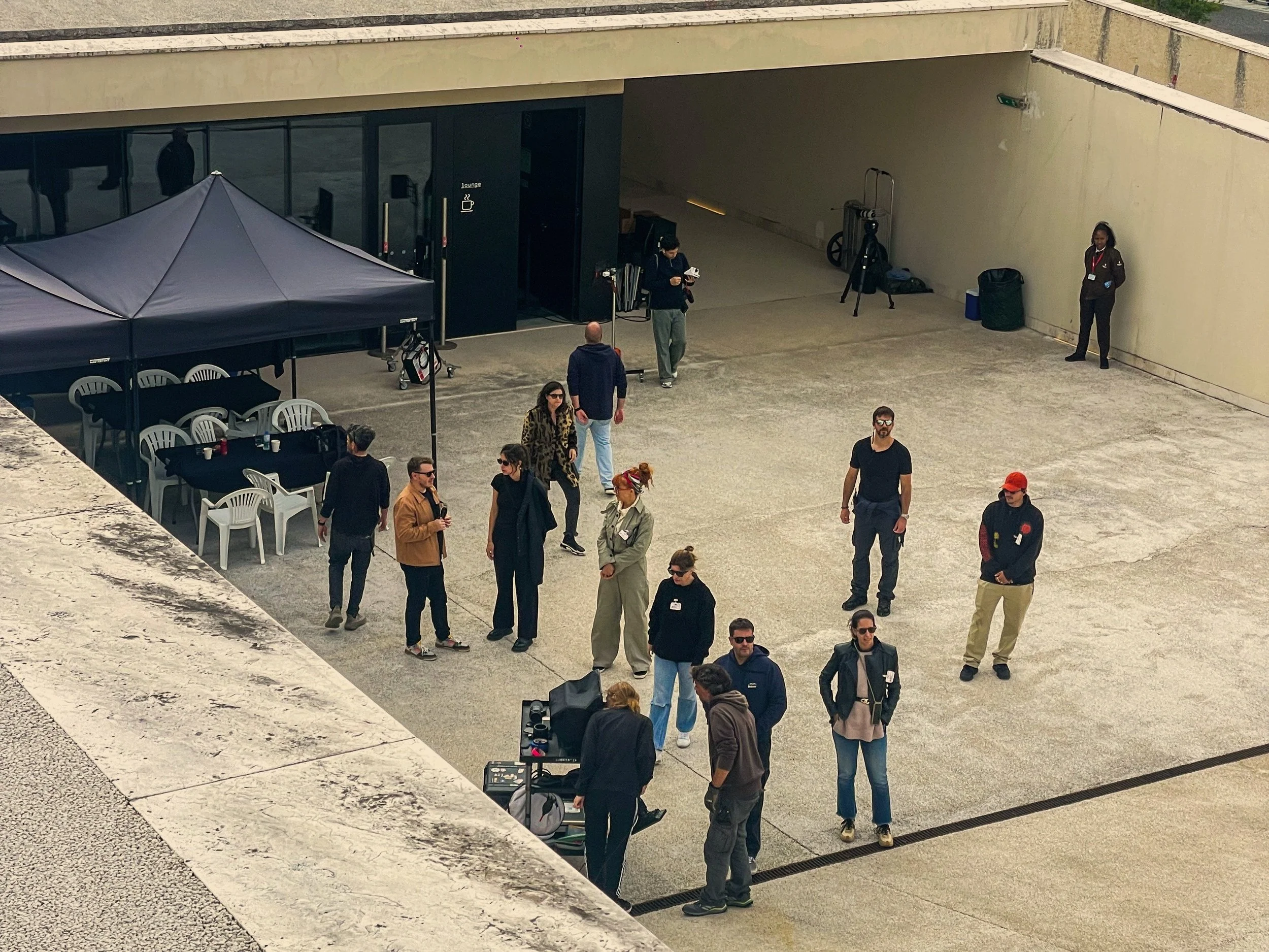 People standing and walking on a rooftop terrace with tables, chairs, and a security guard beside a wall, with equipment set up for a photo or video shoot.
