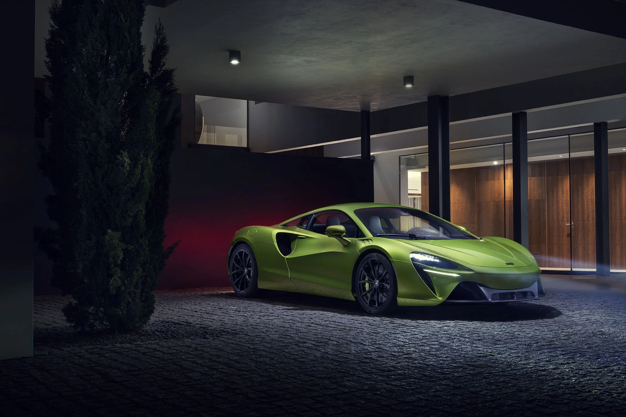 A bright green sports car parked outside a modern house at night, with part of a tree visible on the left and large windows and wooden panels on the house.