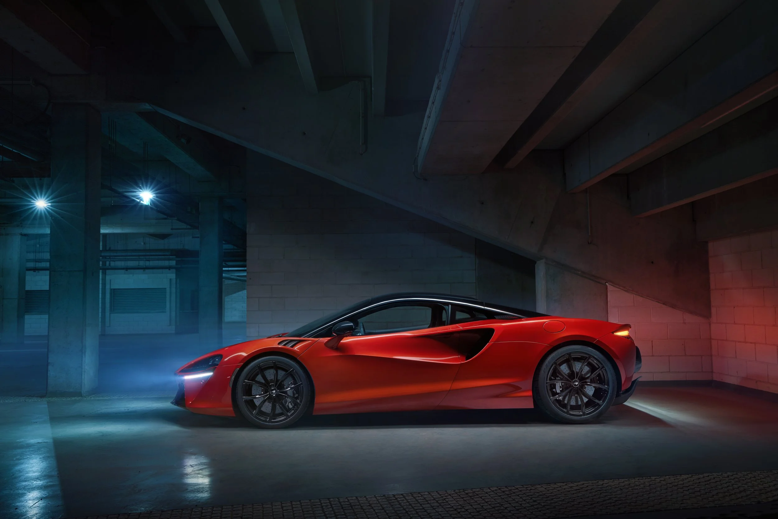 A red sports car parked in a dimly lit underground parking garage with concrete pillars and walls, illuminated by blue and red lights.