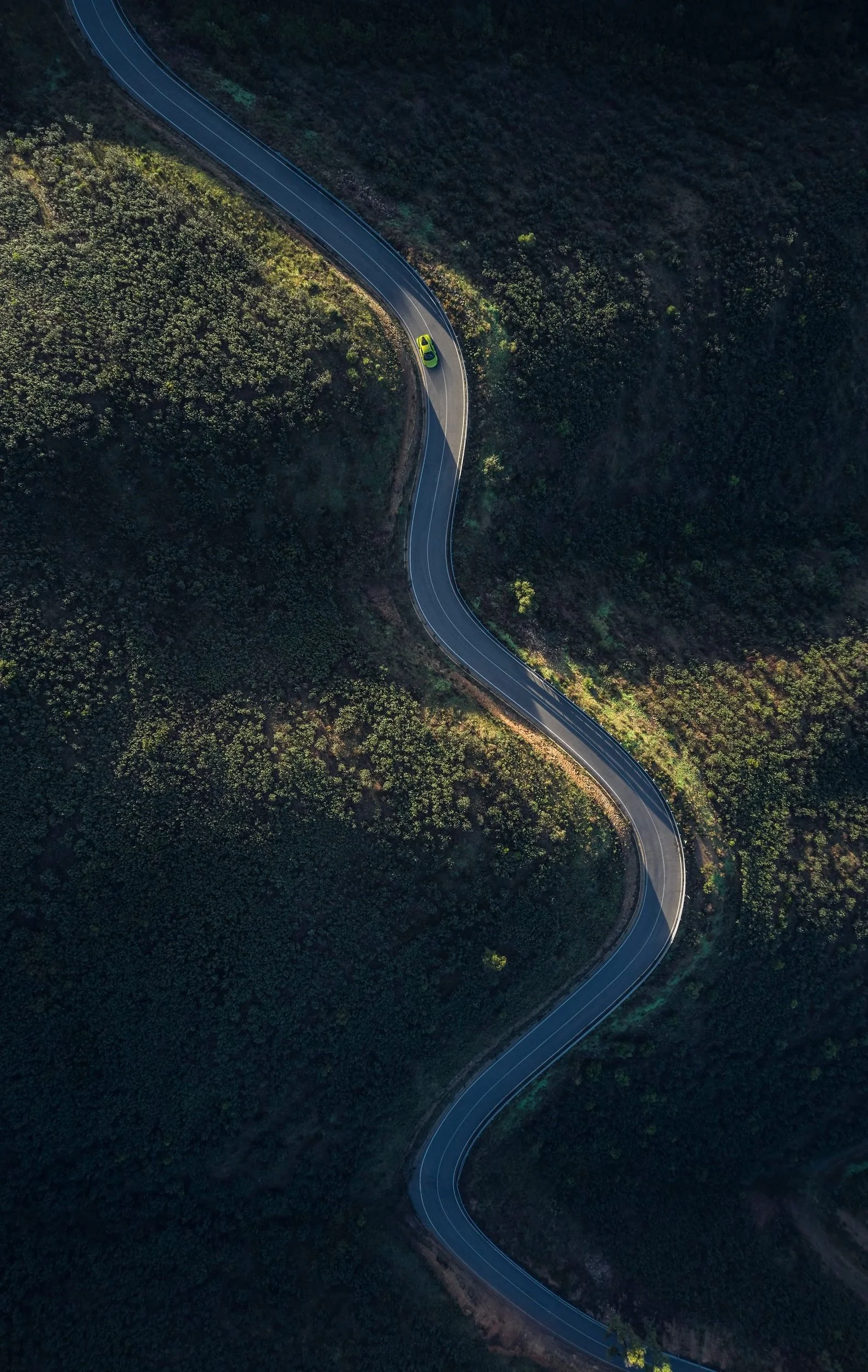 An aerial view of a winding mountain road with a single bright green car traveling through dense forest and hillside landscape.