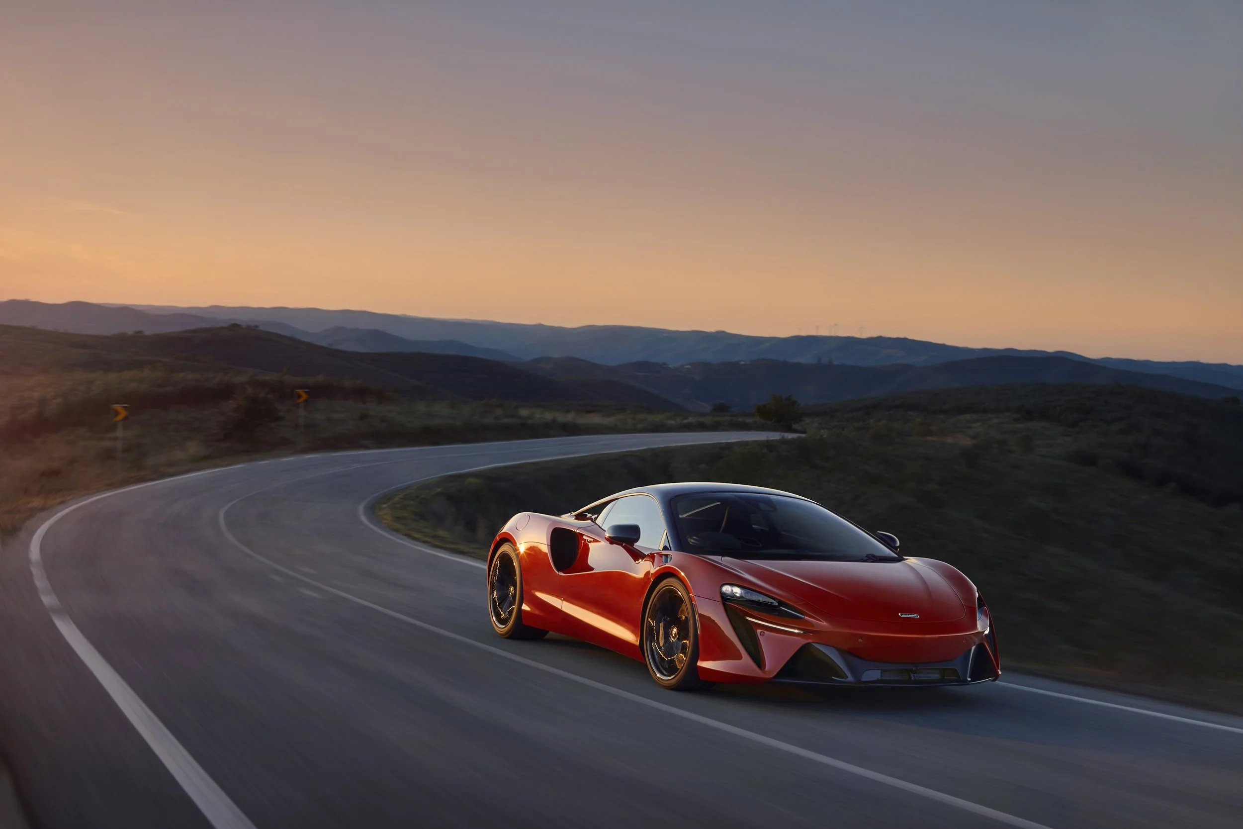 A red sports car driving along a winding mountain road at sunset with scenic hills and mountains in the background.