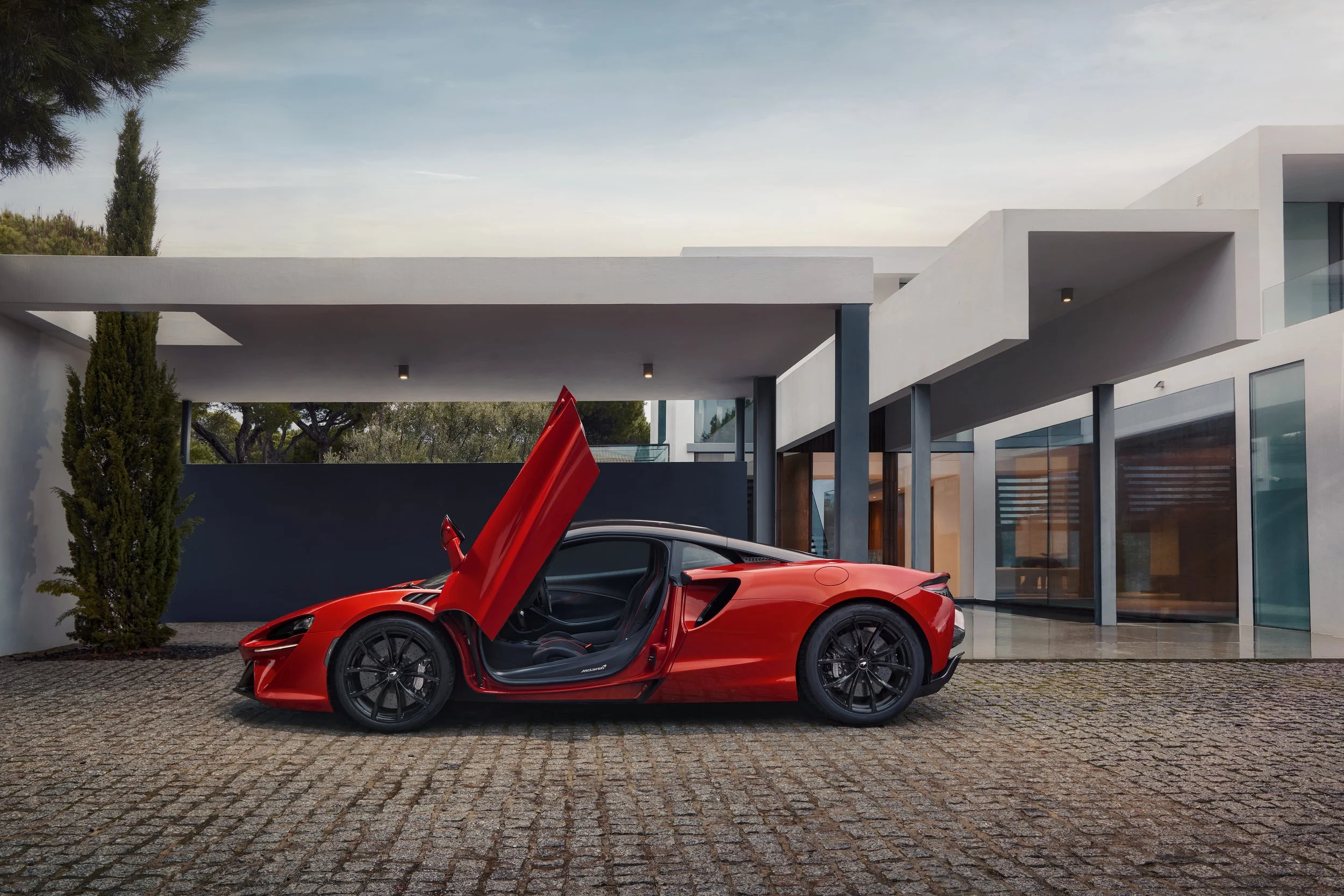 Red sports car with butterfly doors open parked in front of a modern white building with large glass windows and a cobblestone driveway, alongside a tall cypress tree.