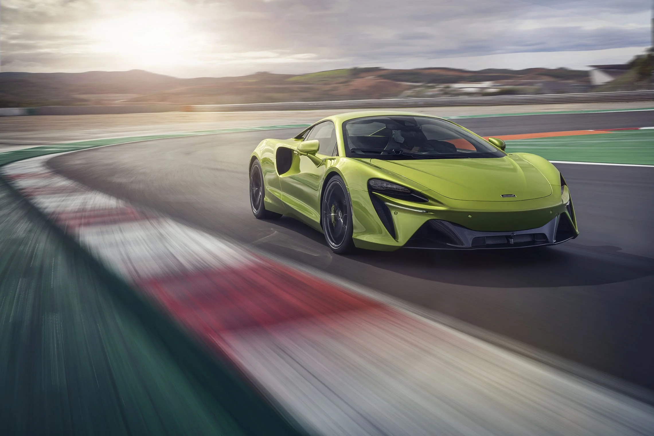 A bright green sports car racing on a curved race track with scenic hills in the background and a cloudy sky overhead.
