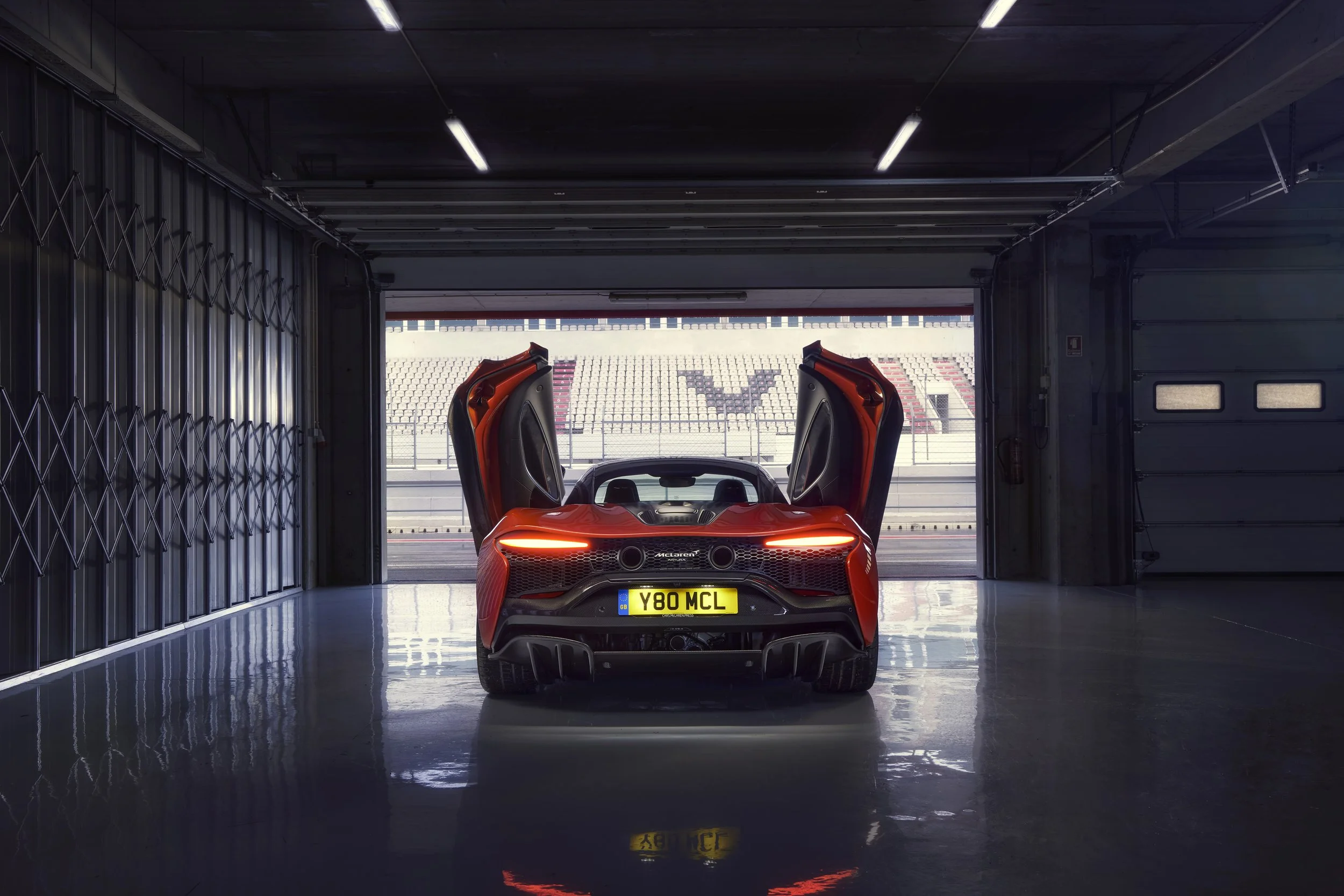 A red McLaren sports car with butterfly doors open, parked inside a garage with a view of a race track and empty grandstands outside.