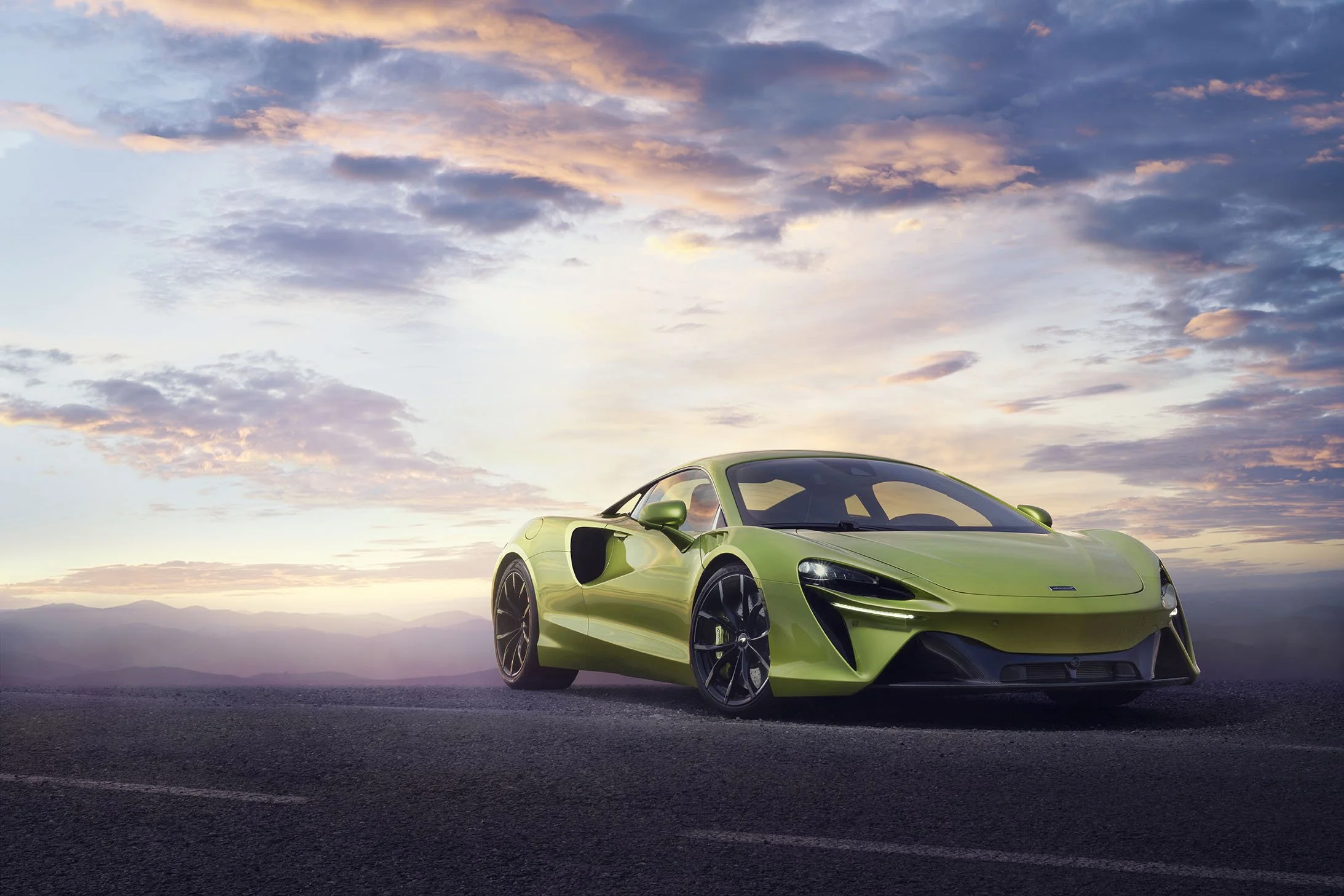 Green sports car parked on a dark road with a sunset sky and clouds in the background.