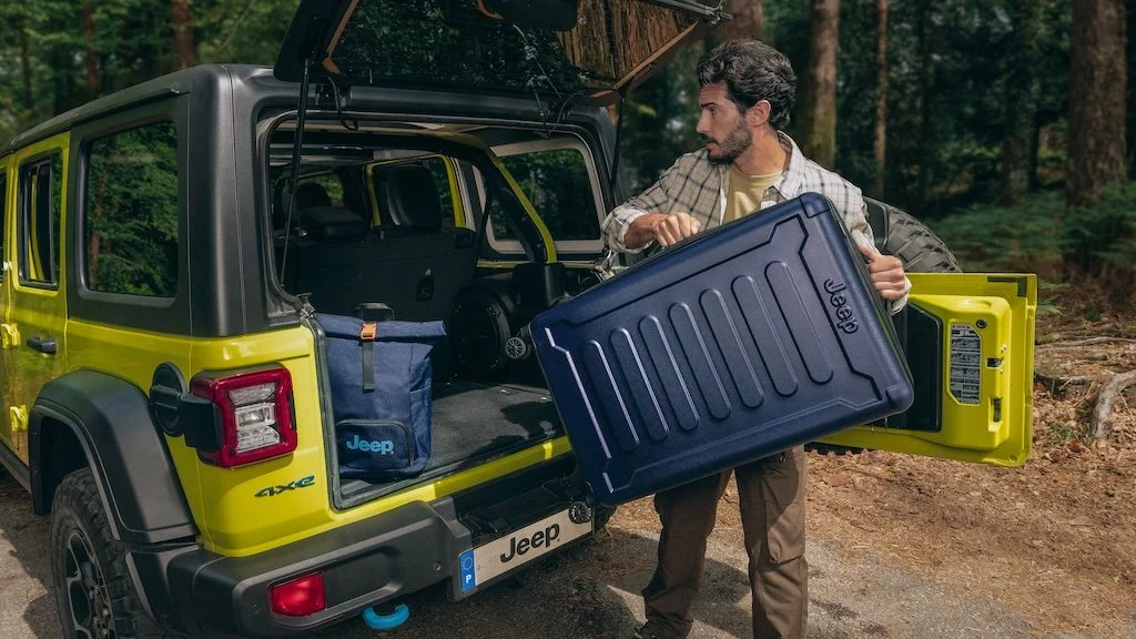 Man loading a large blue cooler into the back of a yellow Jeep in a forested area.