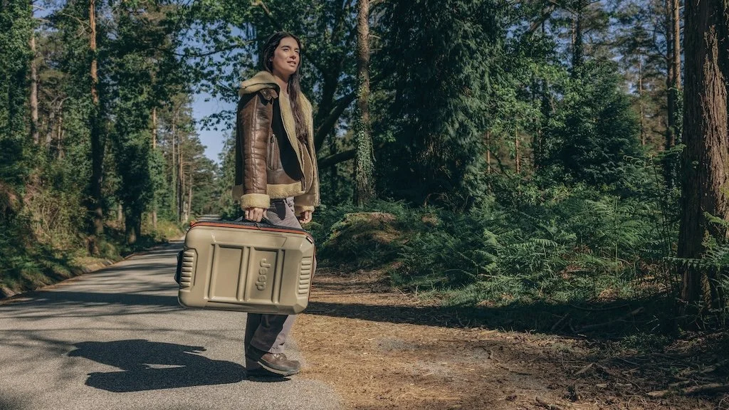 A woman standing on a forest road holding a beige hard case with a Jeep logo, wearing a brown leather jacket with shearling lining, in a wooded area with tall trees and sunlight.