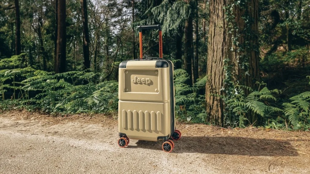 A beige suitcase with black handles and orange wheels standing upright on a dirt path in a forest, casting a shadow.