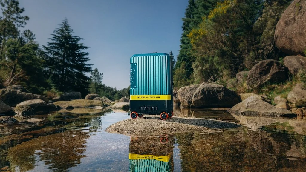 A small, blue suitcase with black and red wheels stands on a rock in a shallow river, surrounded by rocks and trees under a clear sky.