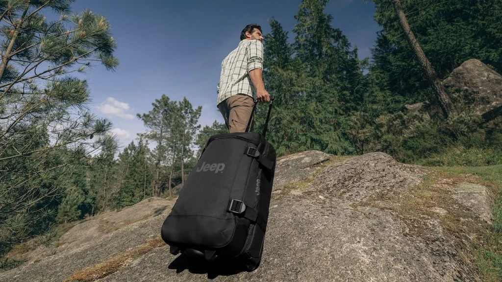 Person standing on large rock in forest, pulling a black rolling bag with Jeep logo.