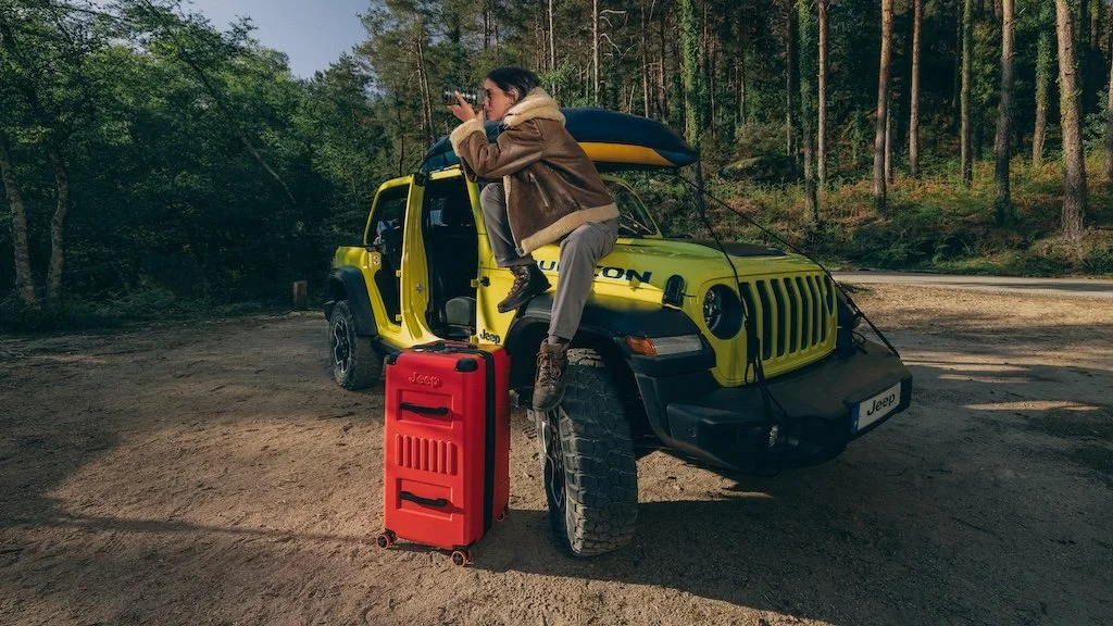 A person is sitting on the hood of a yellow Jeep vehicle, taking a photograph or looking through binoculars, with a red suitcase on the ground next to it. The scene is set in a wooded outdoor area.