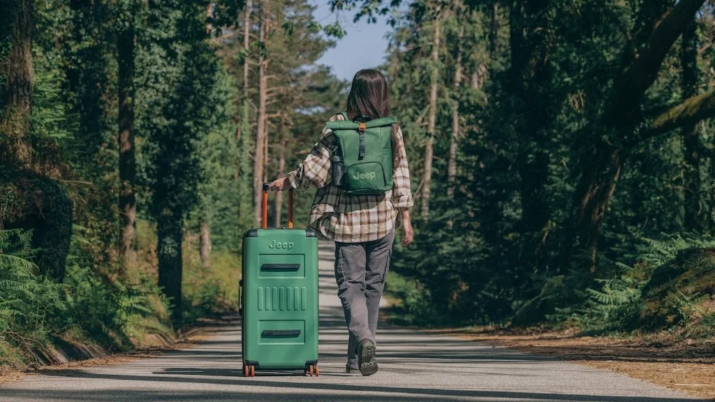 Woman walking down a forest road pulling a green suitcase with a green backpack, surrounded by tall trees.