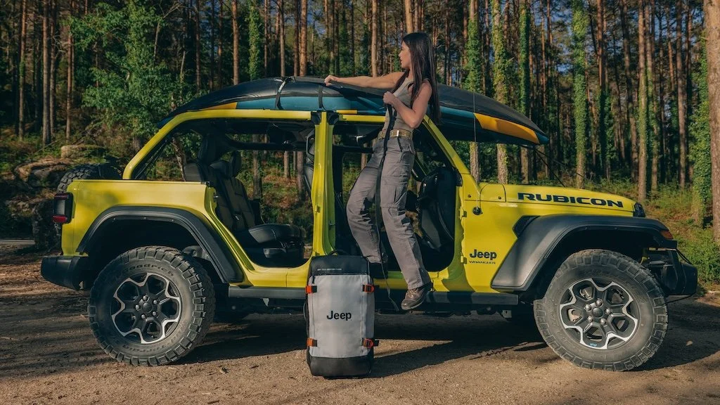 A woman standing on the side of a yellow Jeep Rubicon, holding a paddle, with a backpack on the ground nearby, in a forest setting.