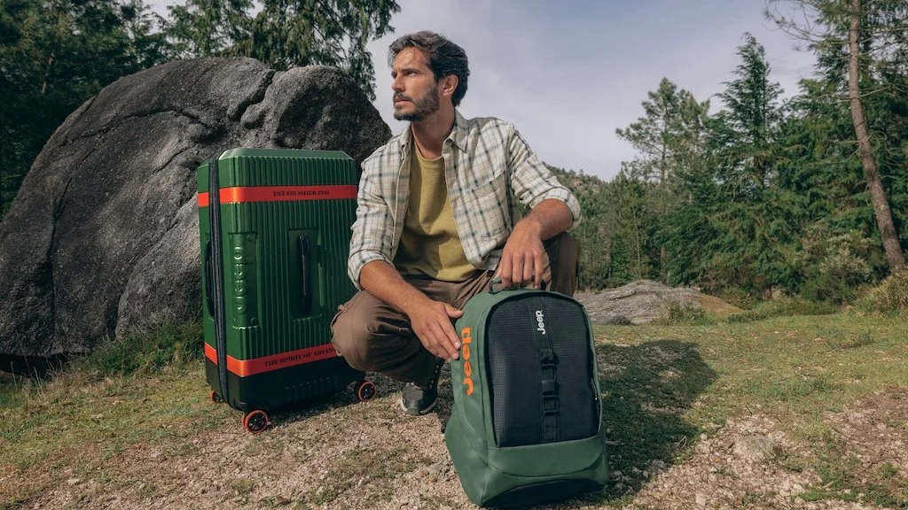 A man with a beard and casual clothing sitting outdoors surrounded by trees and rocks, with a large green suitcase with red accents and a smaller green and black backpack.
