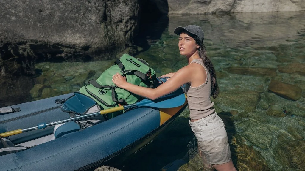 A woman in tank top, shorts, and a cap stands in water next to a kayak, holding a paddle and looking back.