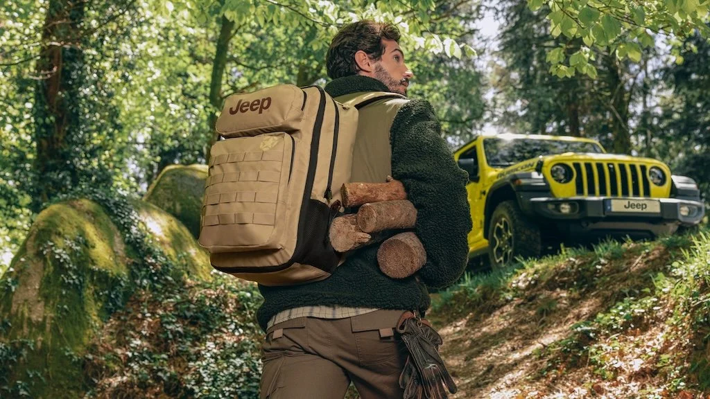 A man in outdoor clothing carrying firewood on his shoulder, with a yellow Jeep parked in the forest behind him.
