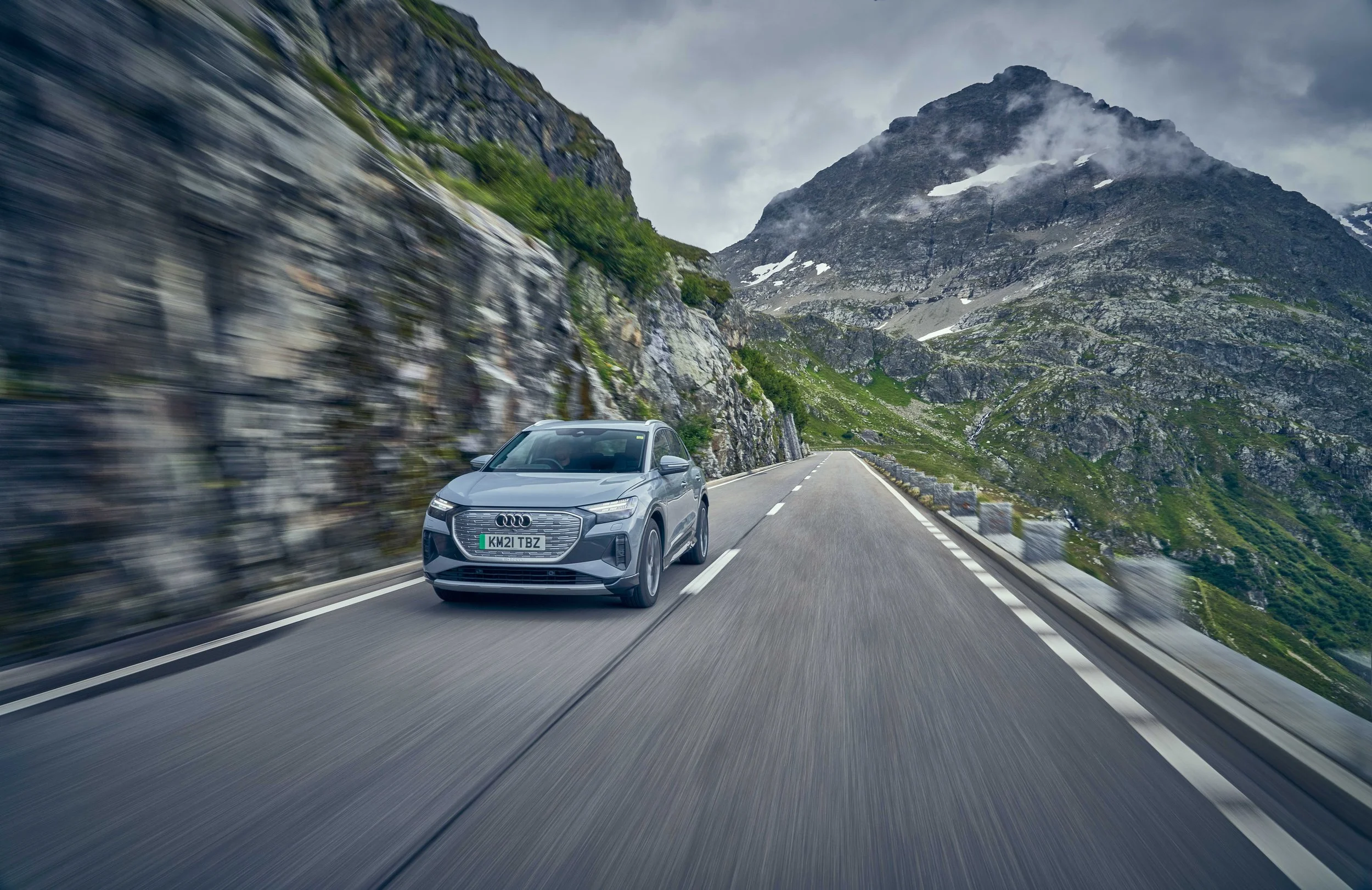 A silver Audi SUV driving on a mountain road with rocky terrain and green vegetation, with mountains and cloudy sky in the background.