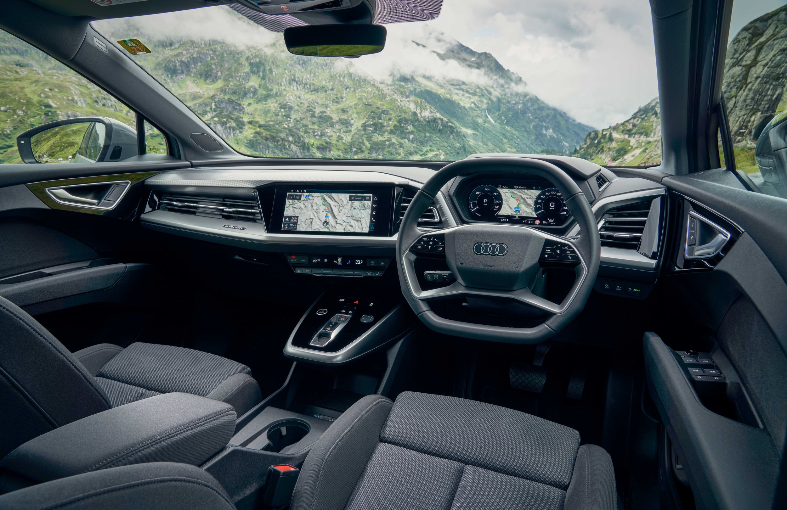 The image captures the interior of a modern car, showing the dashboard, steering wheel, and digital instrument cluster. Through the windshield, a scenic view of green mountains and cloudy skies is visible.