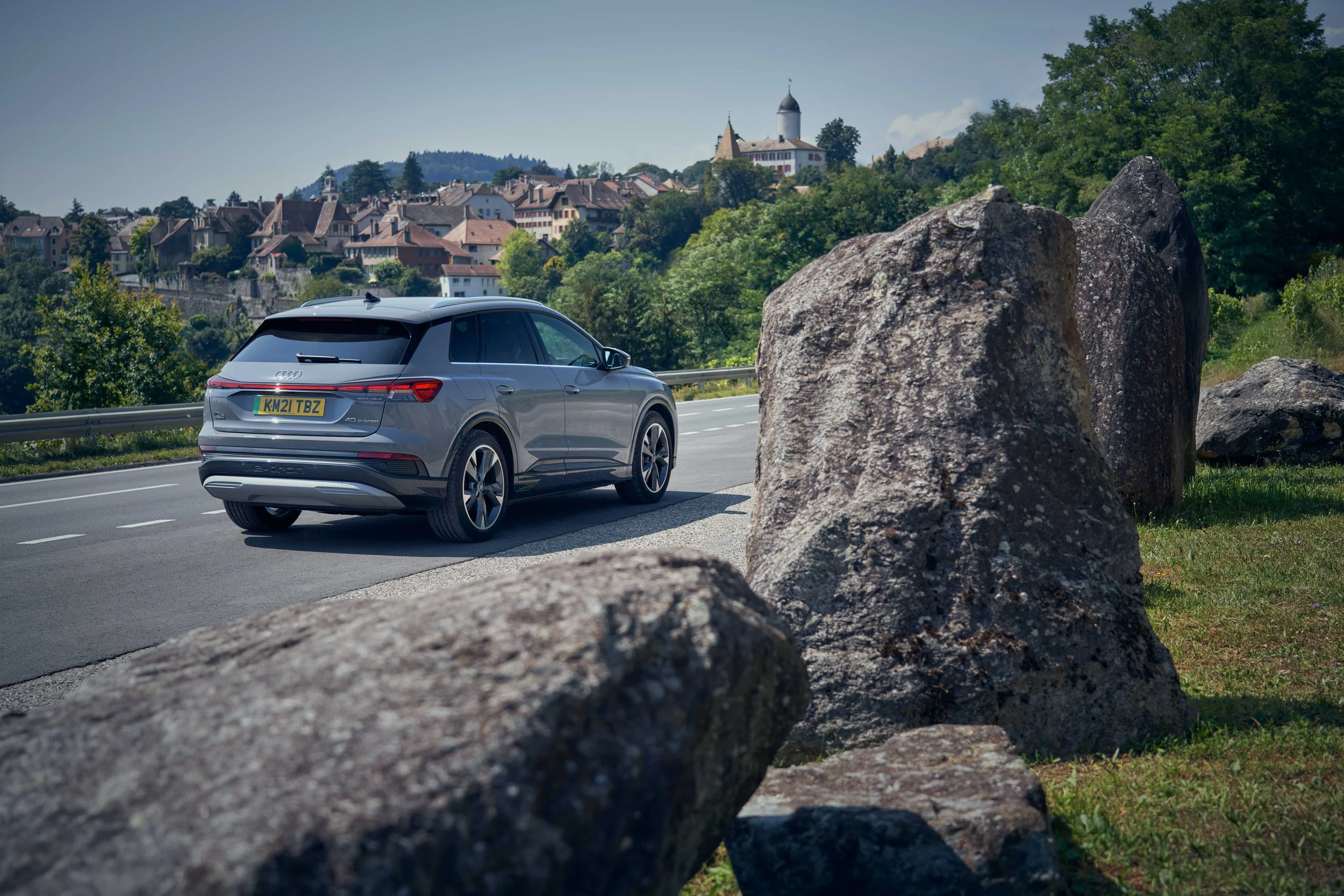 A modern silver SUV driving along a scenic highway with a small town and castle in the background, partially obscured by large rocks near the roadside.
