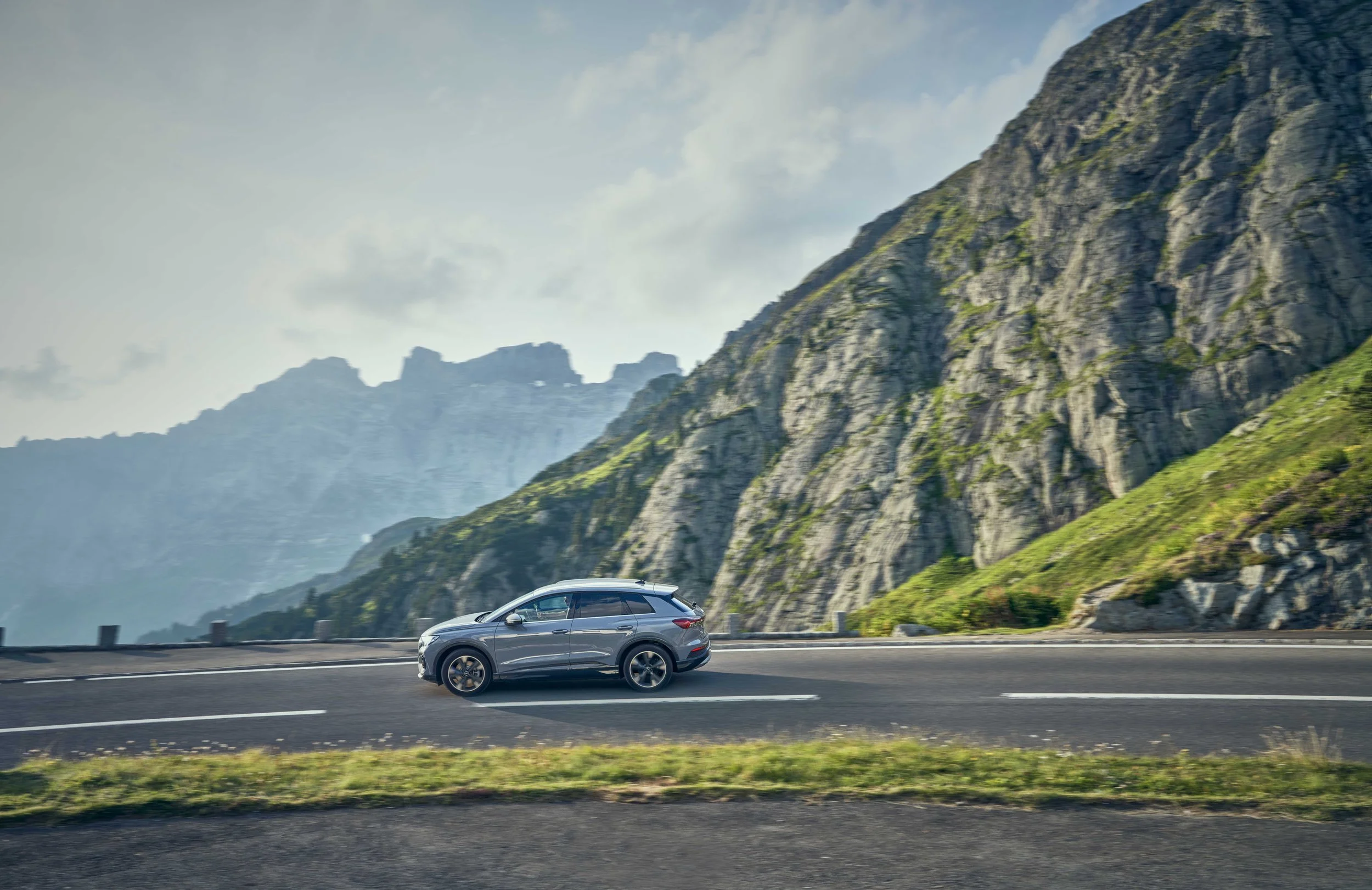 A gray SUV driving on a mountain highway with steep, rocky, and green-covered slopes in the background under a partly cloudy sky.