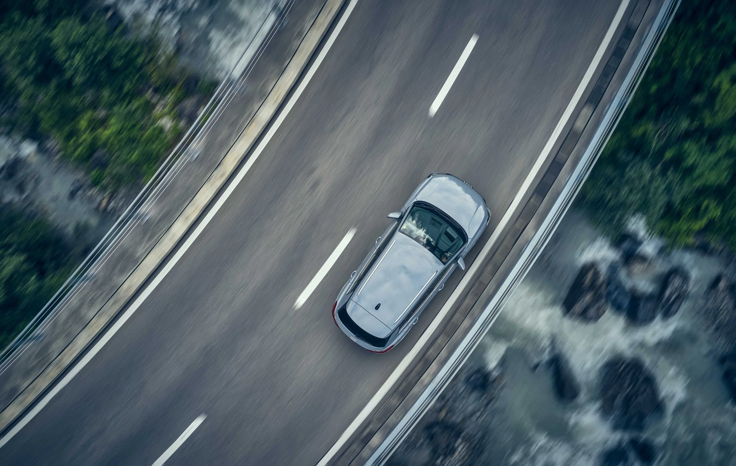 An aerial view of a silver car driving on an open highway surrounded by greenery.