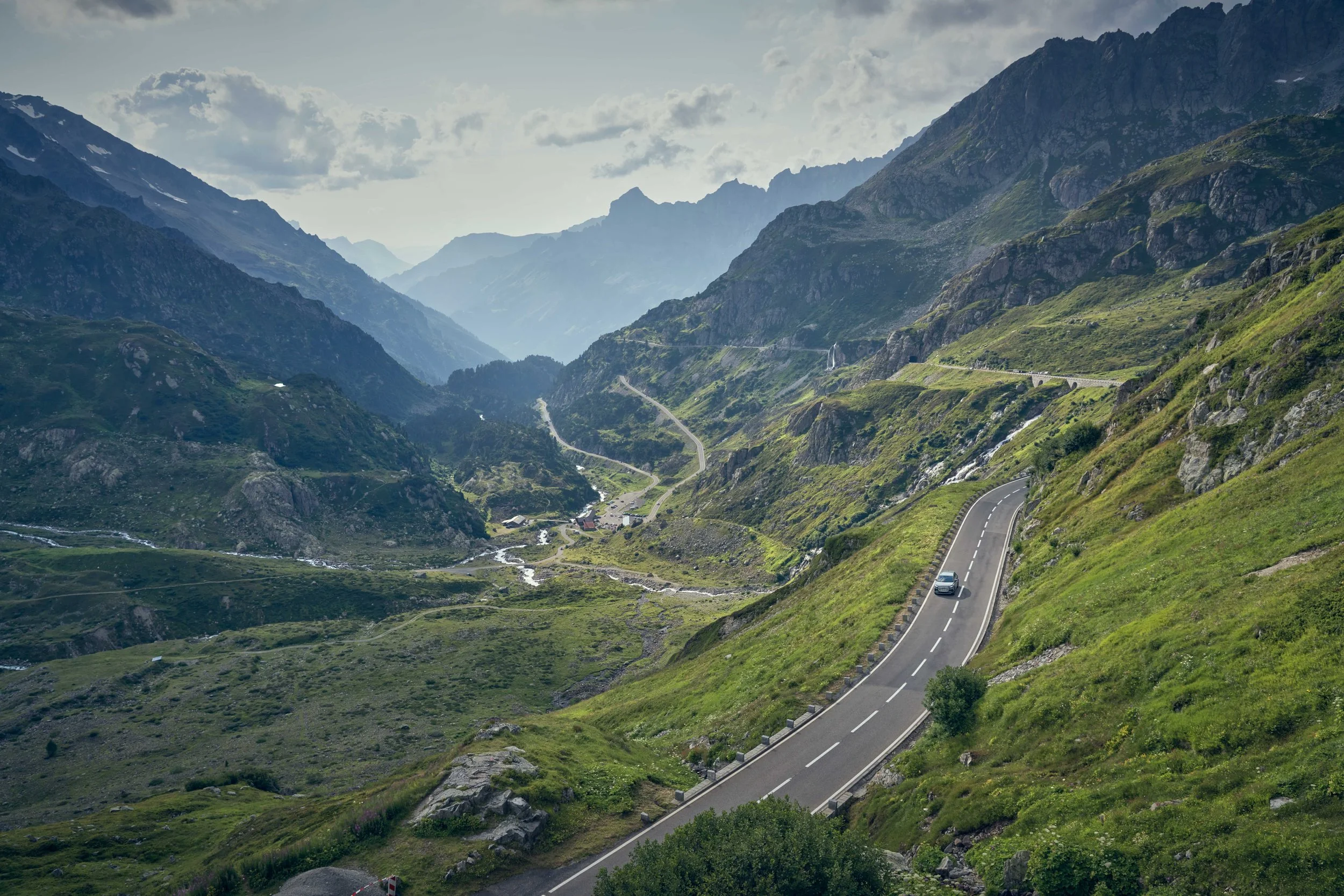 A winding mountain road with a car driving along it, surrounded by lush green hills and towering mountains with patches of snow in the distance, under a partly cloudy sky.