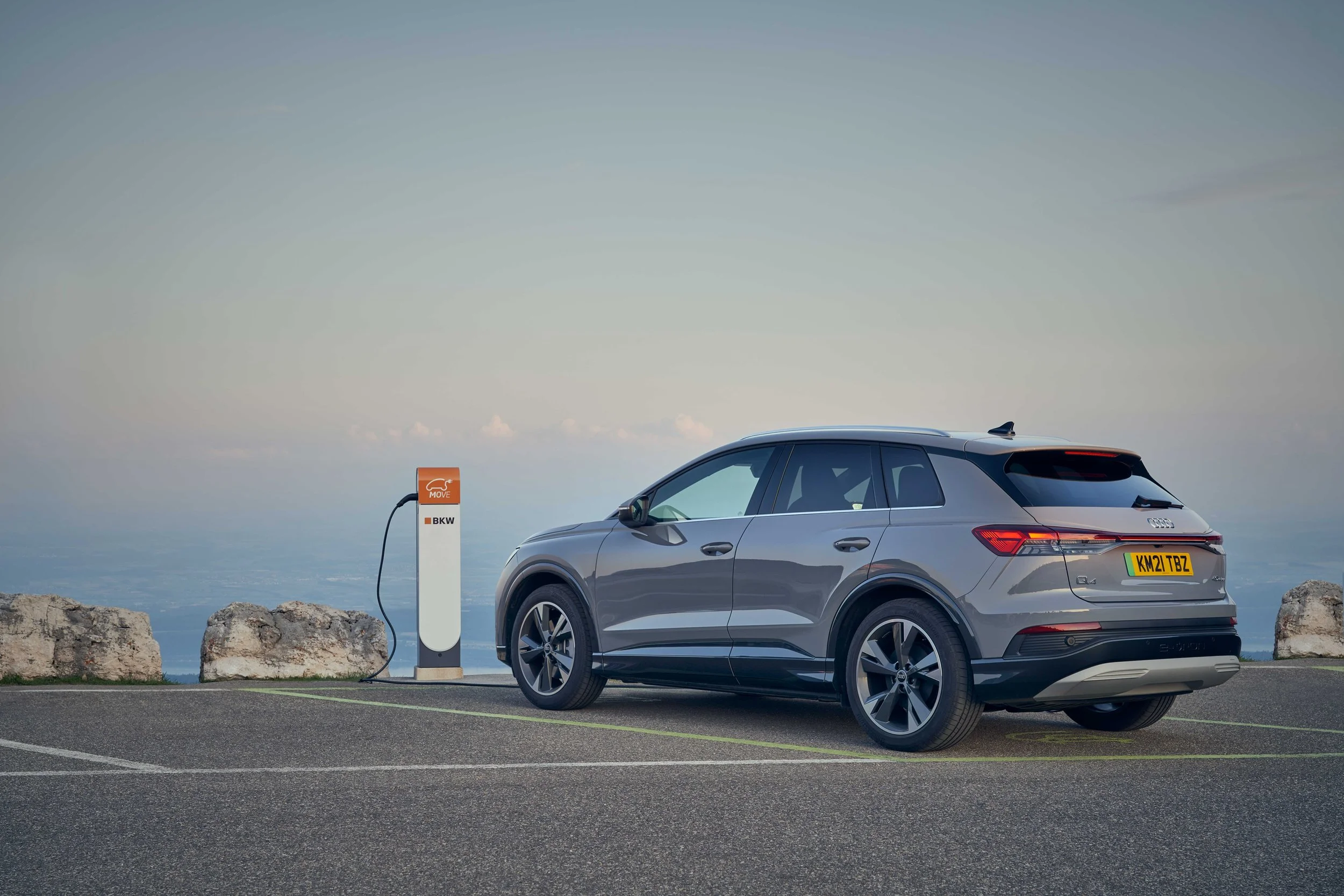 A gray SUV parked at an electric vehicle charging station on a cliff top with rocks and the ocean in the background.