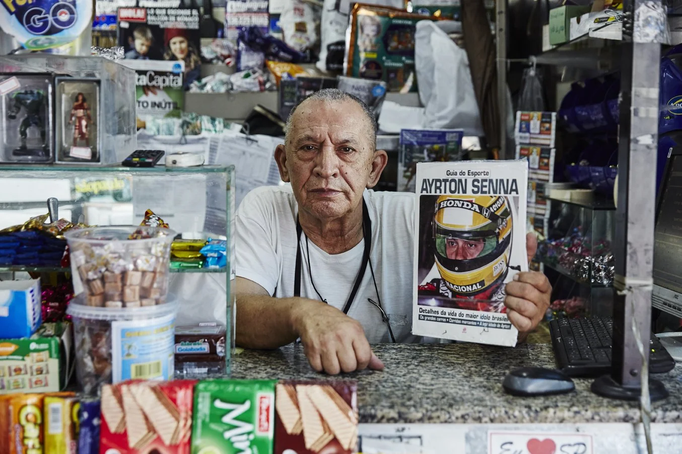 An older man sitting behind a store counter holding a magazine with Ayrton Senna's picture on the cover. The counter has various candies and snacks, and the background is filled with various items and products.