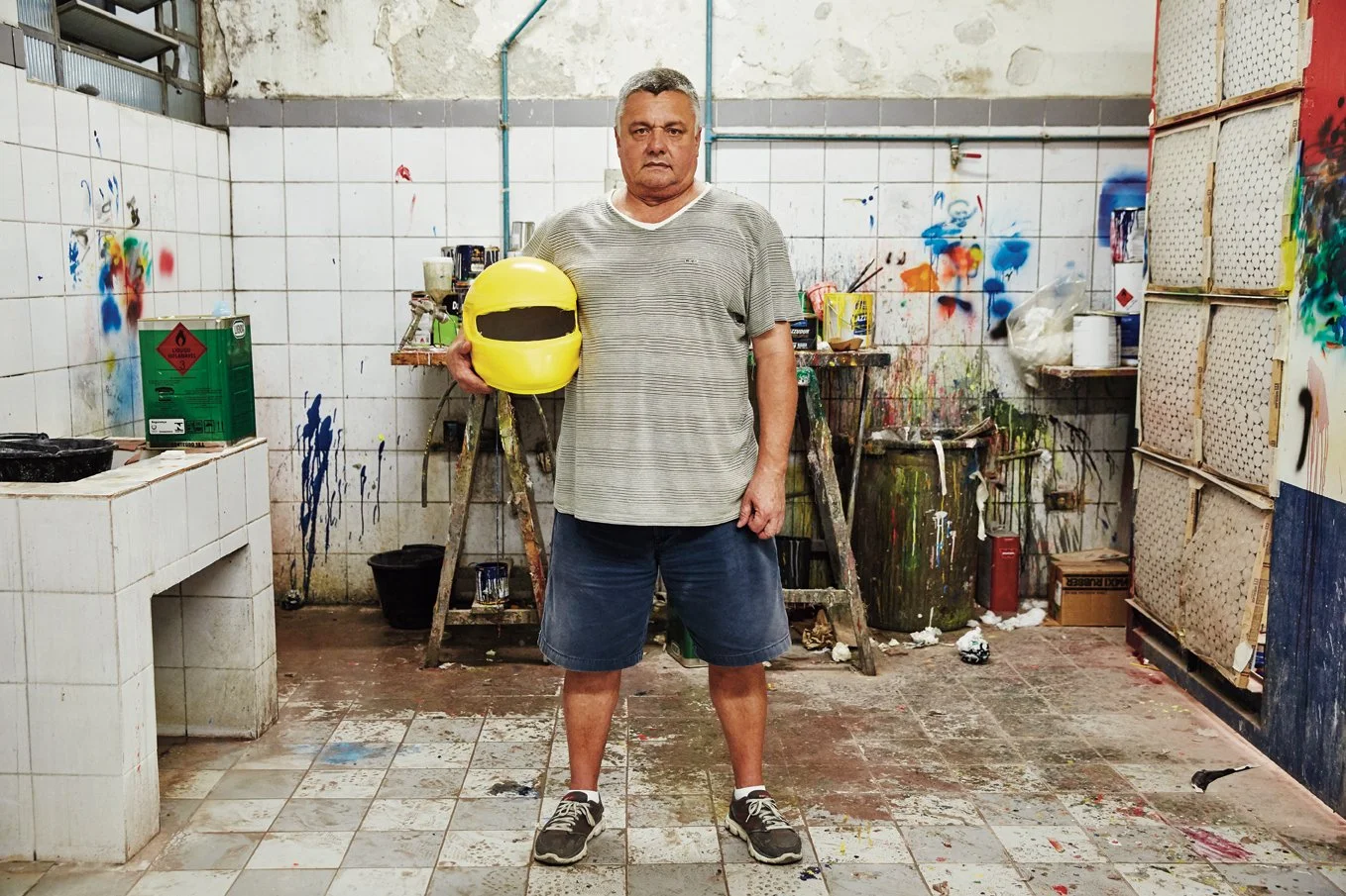 A man holding a yellow helmet standing in a paint-stained art studio or workshop with paint splatters and supplies around him.