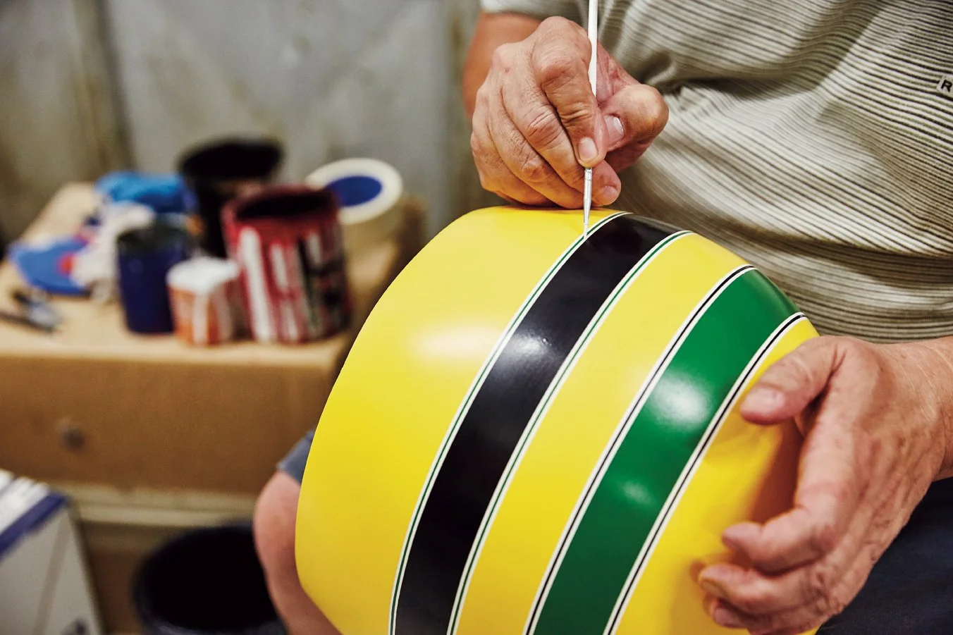 Person painting curved yellow, black, and green stripes on a motorcycle fuel tank with a fine brush.