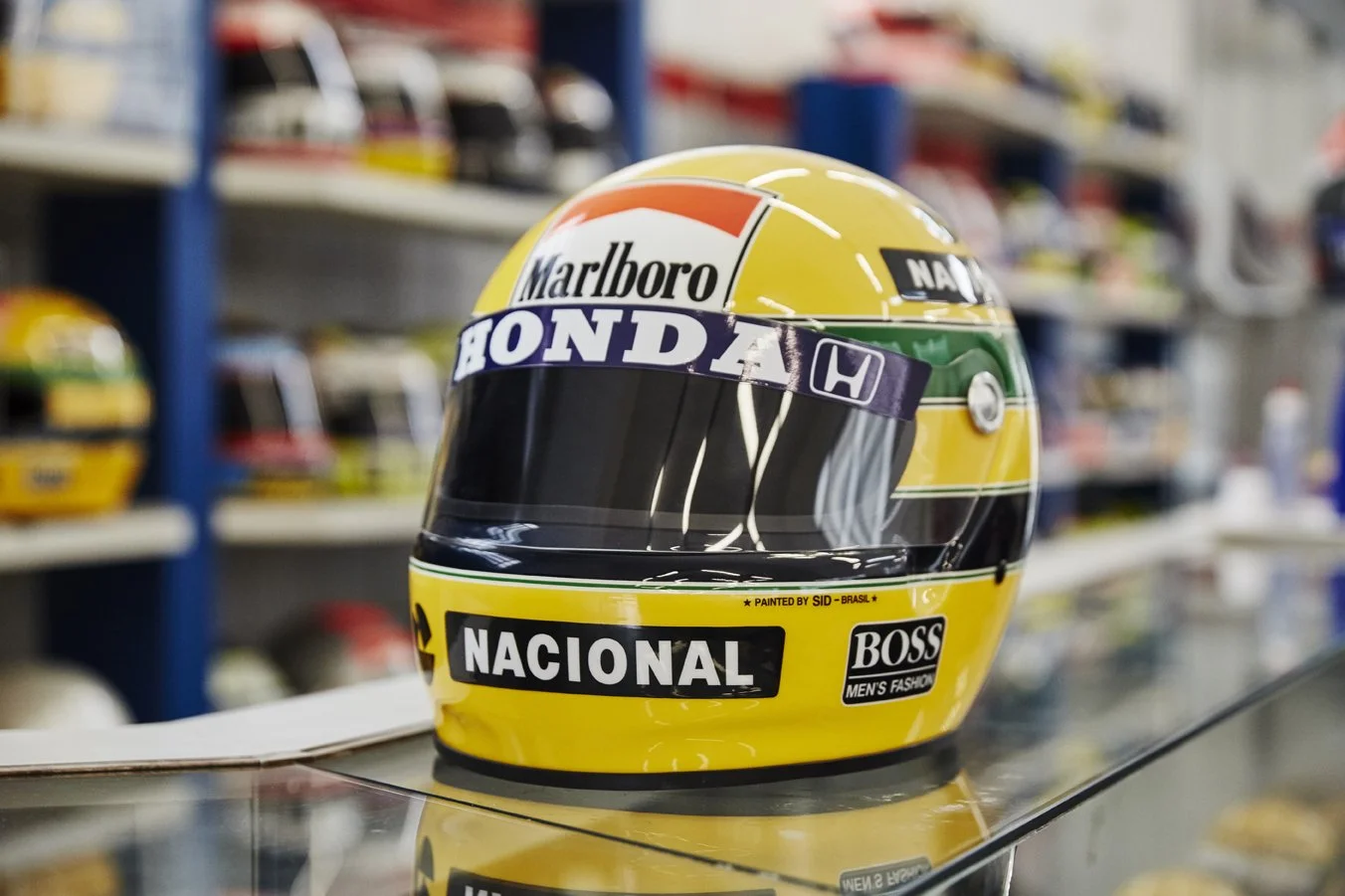 A racing helmet with yellow, black, green, red, and white colors and sponsors' logos, placed on a glass surface in a store with helmets on shelves in the background.
