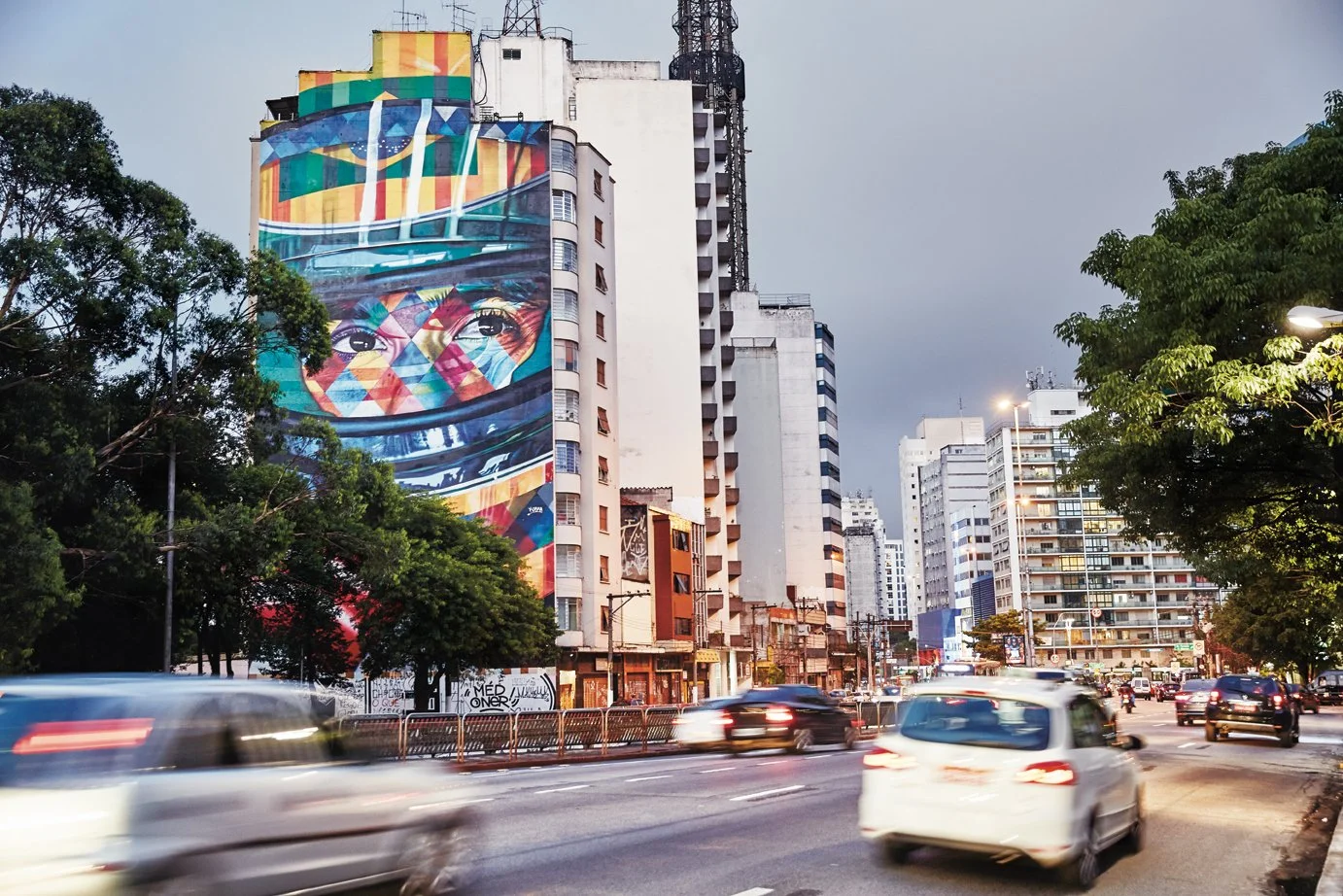 A city street scene at dusk with high-rise buildings, some with colorful murals, and cars moving on the road. There are trees lining the street and streetlights turned on.
