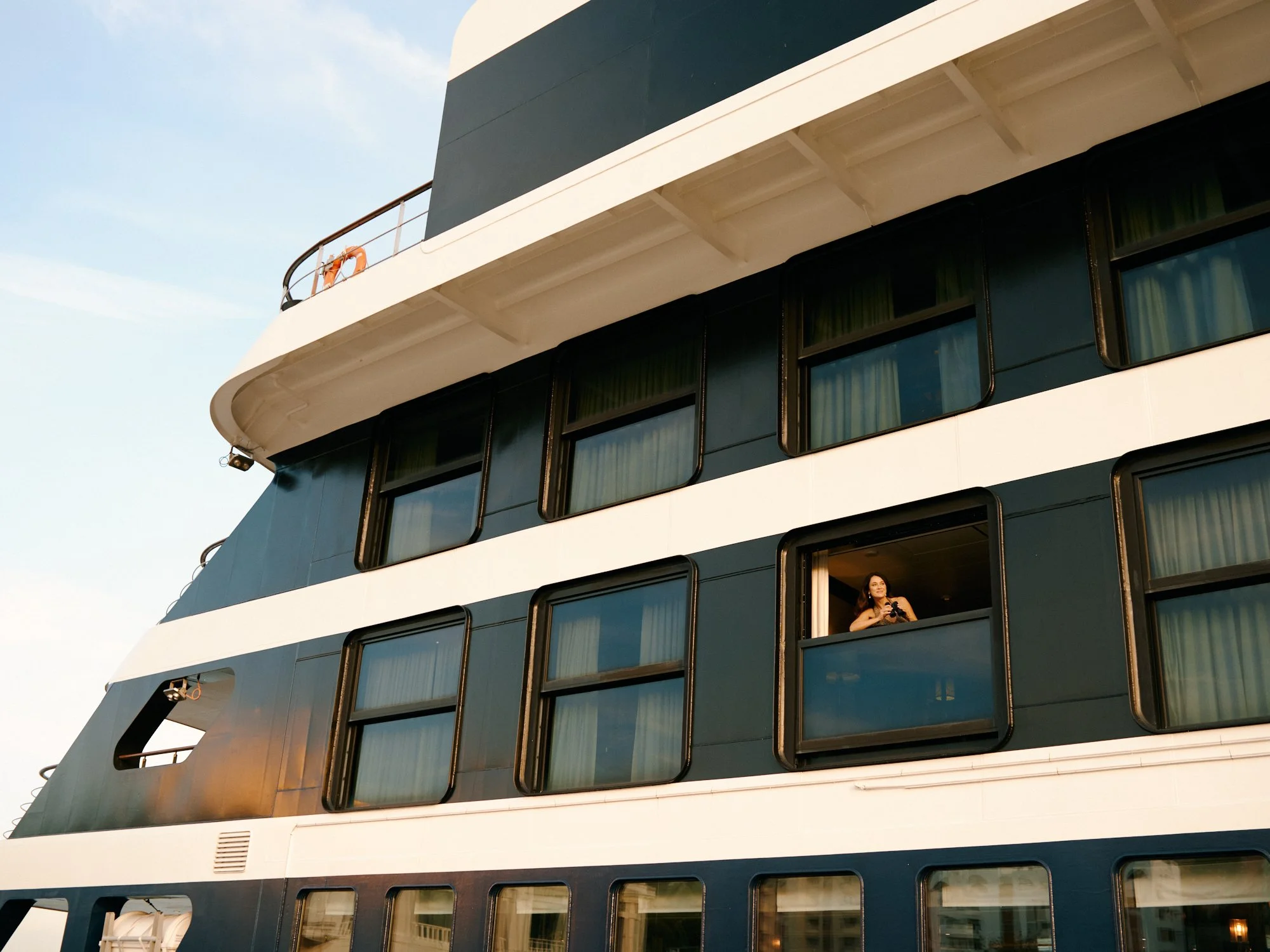 A modern cruise ship with multiple decks featuring large windows and balconies. One woman with long hair is standing at an open window on one of the lower decks, looking out and holding a camera.