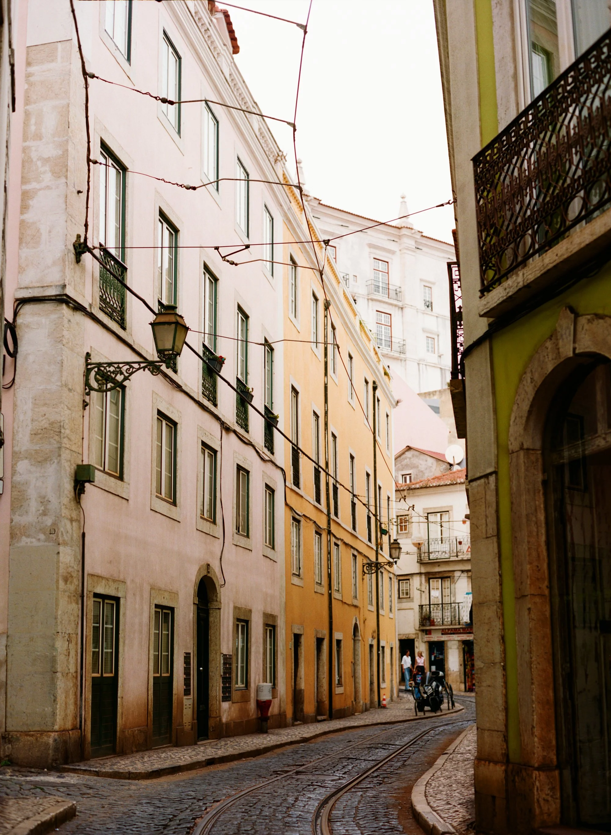 A narrow cobblestone street with tram tracks lined by colorful historic buildings, some with wrought iron balconies, and a street lamp, in a European city.