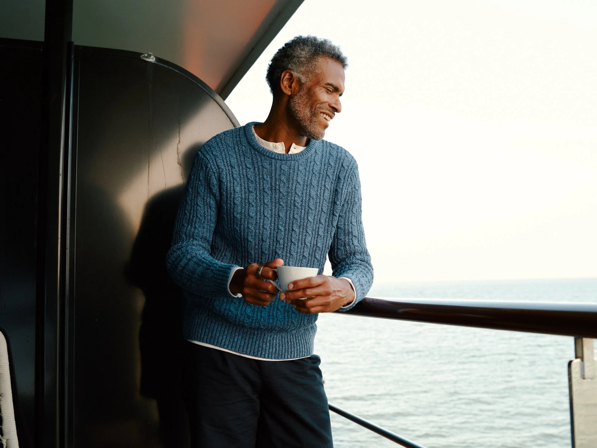 A man with gray hair and a beard is standing on a boat, leaning against the railing, holding a mug, and smiling while looking out at the ocean.