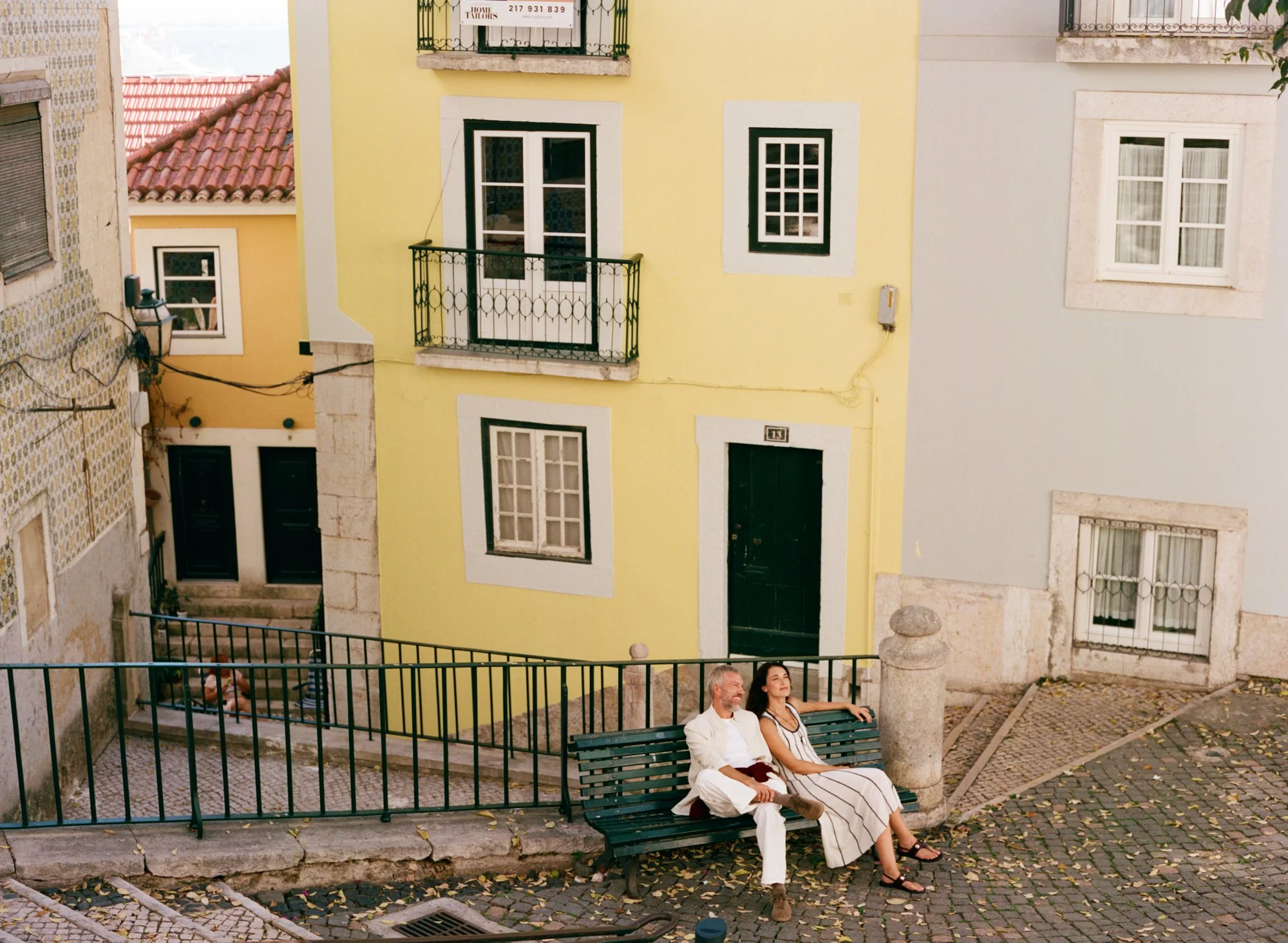 A man and woman sitting on a green park bench in a European city, with colorful old buildings and cobblestone streets in the background.