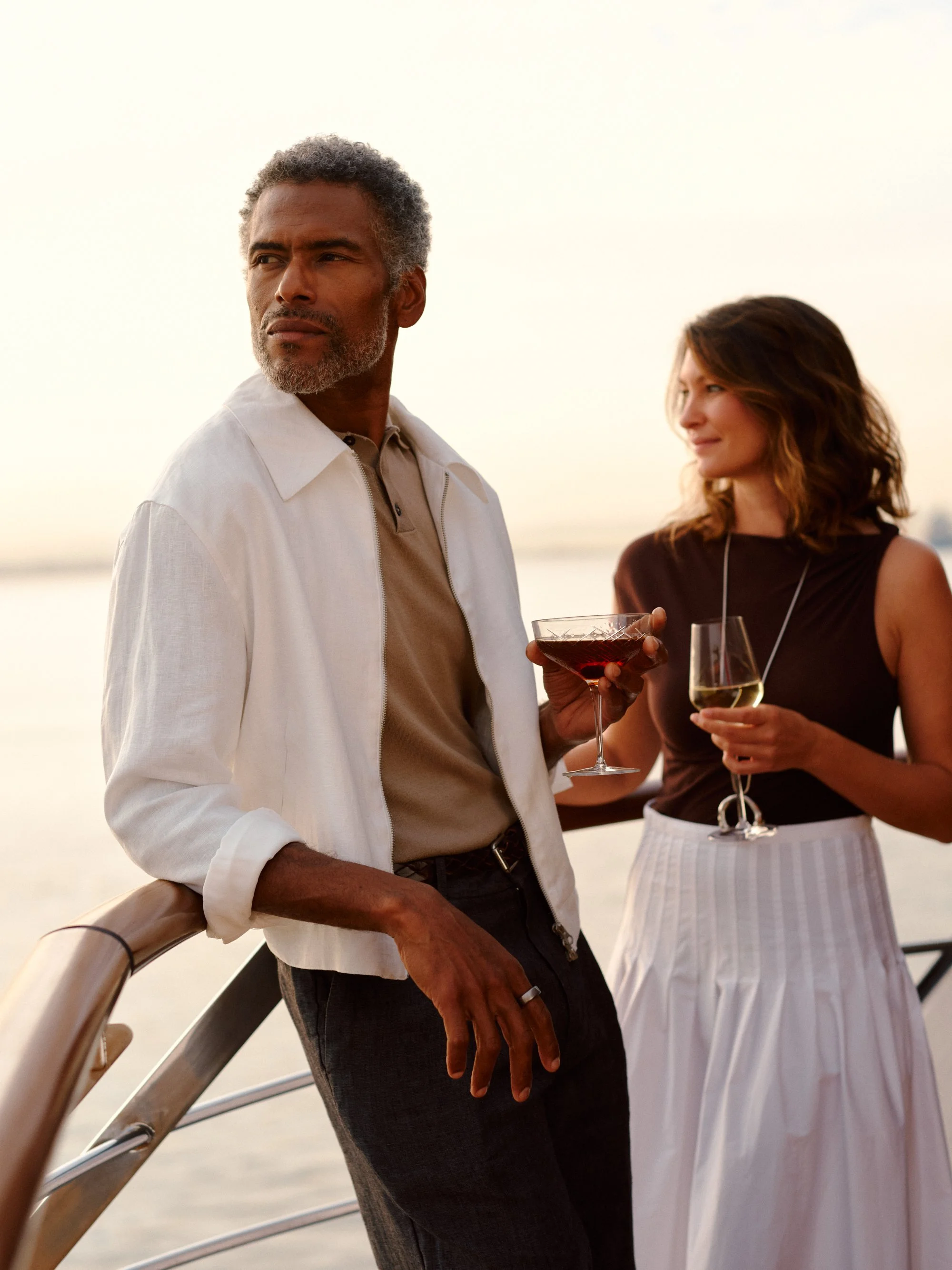 A man and a woman are standing outdoors, holding wine glasses and talking. The man has gray hair and a beard, and is wearing a white shirt and dark pants. The woman has shoulder-length brown hair, and is wearing a dark top and a white striped skirt. The background shows a body of water and a sunset sky.