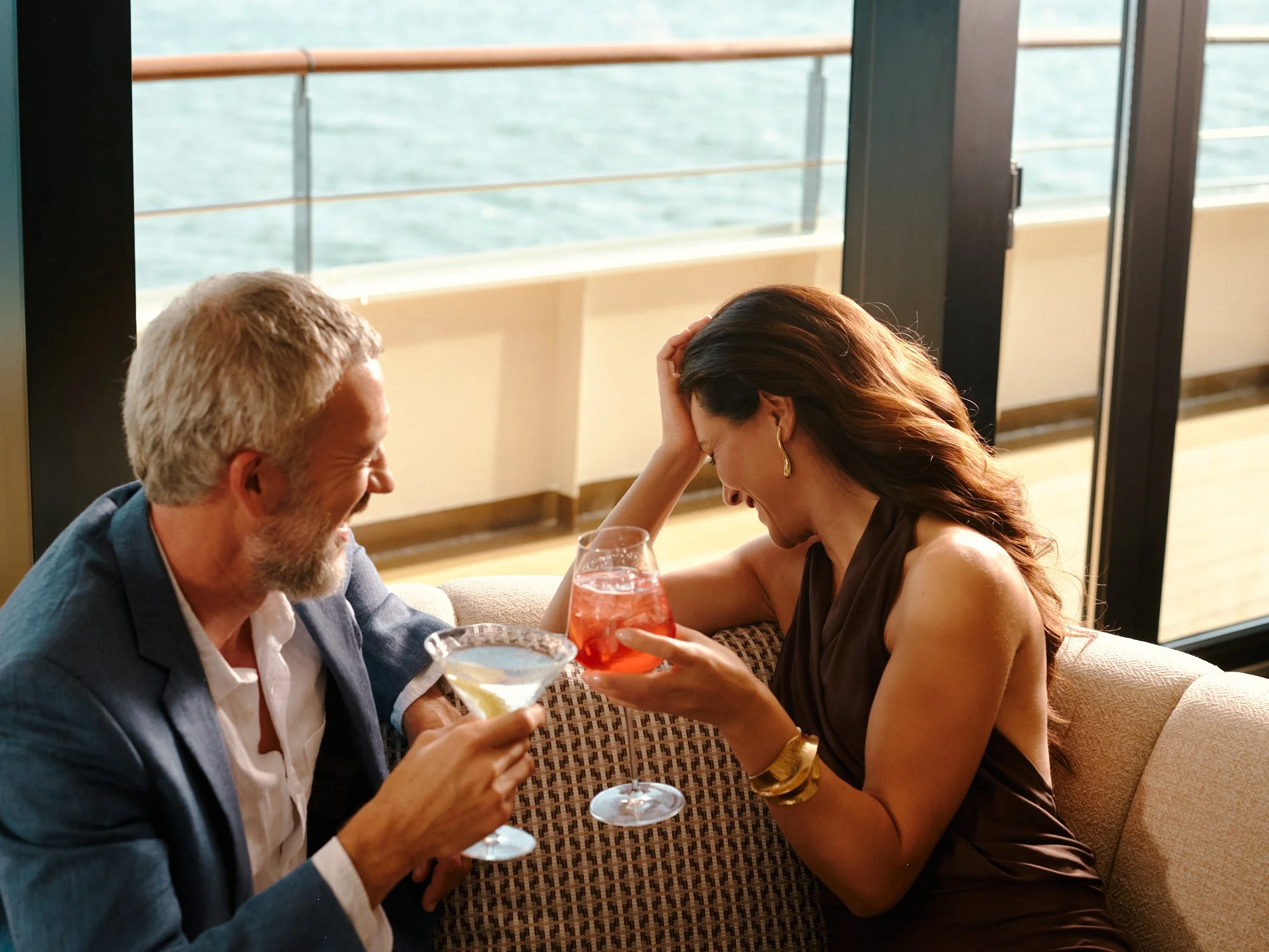 A man and a woman sitting together on a boat, sharing a moment of laughter and conversation while holding drinks, with a large window showing water in the background.