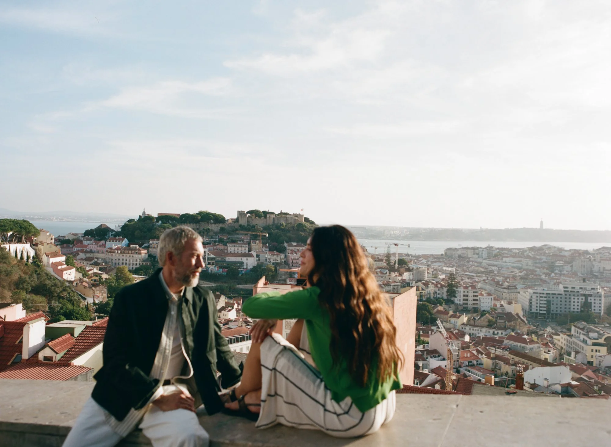 A man and woman sitting on the ground outdoors, talking with a cityscape and a castle on a hill in the background during daytime.