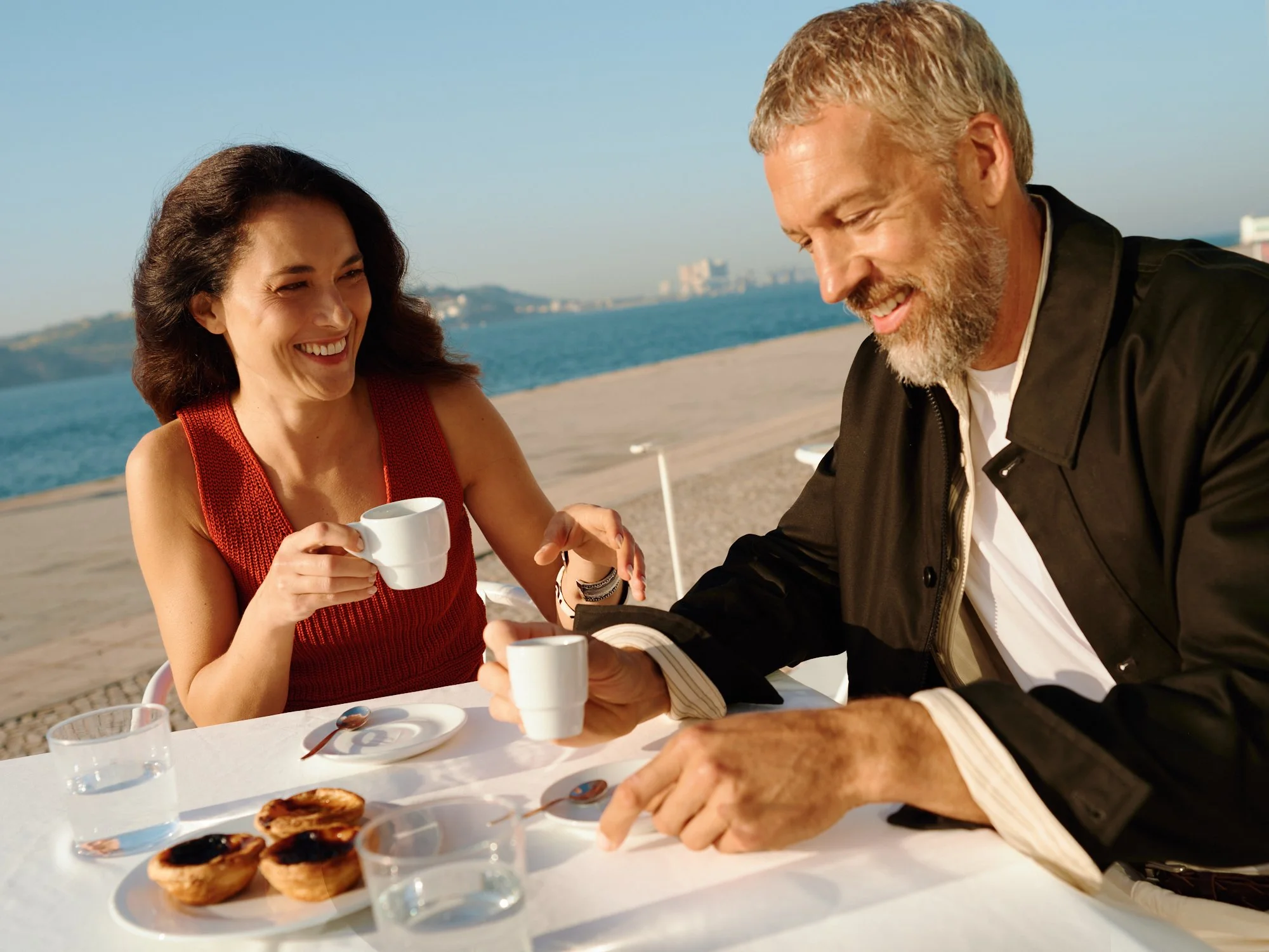Smiling woman in red top and man with gray beard sharing coffee at an outdoor table by the beach with pastries and tea cups in front.