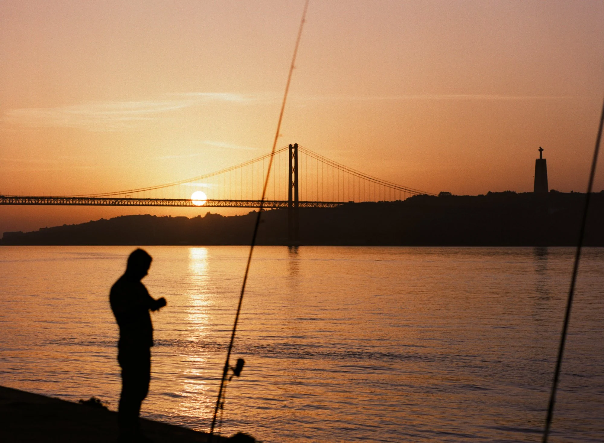 A person standing by the water at sunset with the San Francisco-Oakland Bay Bridge and a lighthouse in the distance.