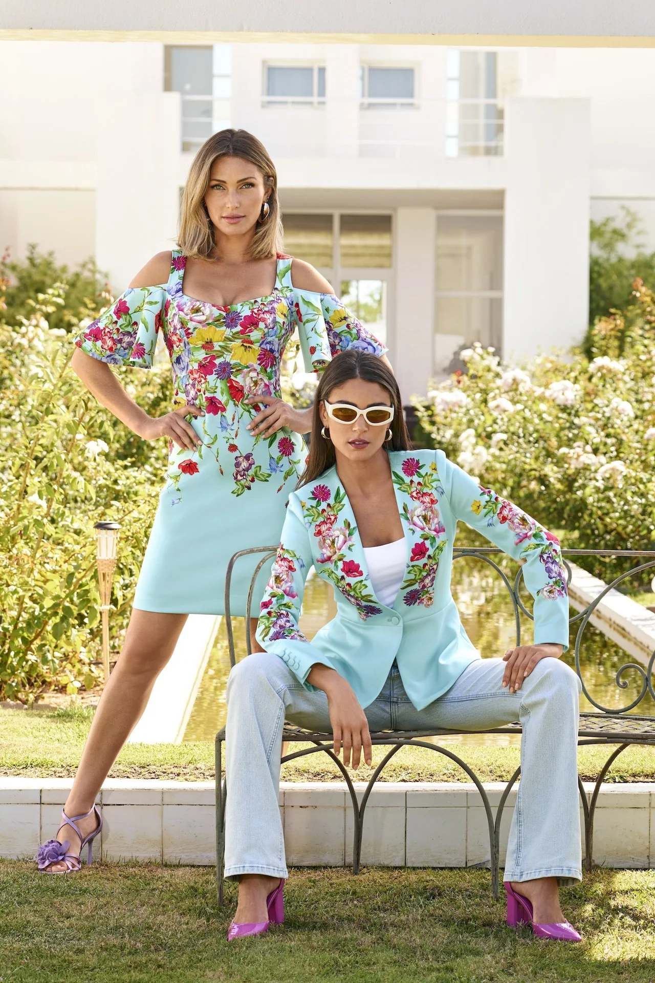 Two women dressed in floral summer outfits pose outdoors near a garden with a modern white building in the background. The woman standing has blonde hair and wears a colorful floral dress with shoulder cutouts. The woman sitting has dark hair, white sunglasses, and wears a light blue floral blazer with white jeans. Both women wear pink shoes.